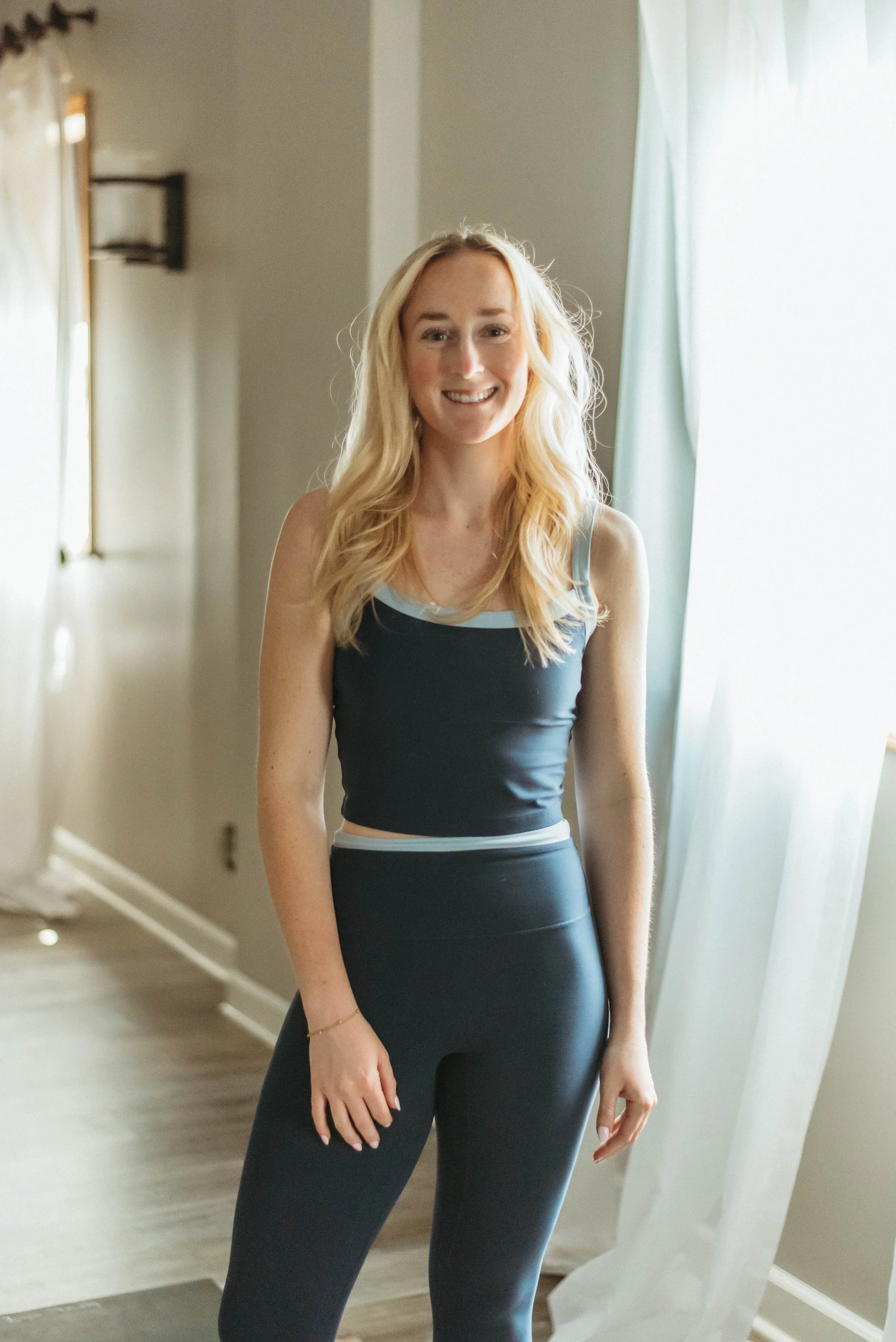 Young woman with brown hair tied back, smiling, wearing a teal sports bra and black leggings, standing near a window with curtains and a candle in the background.