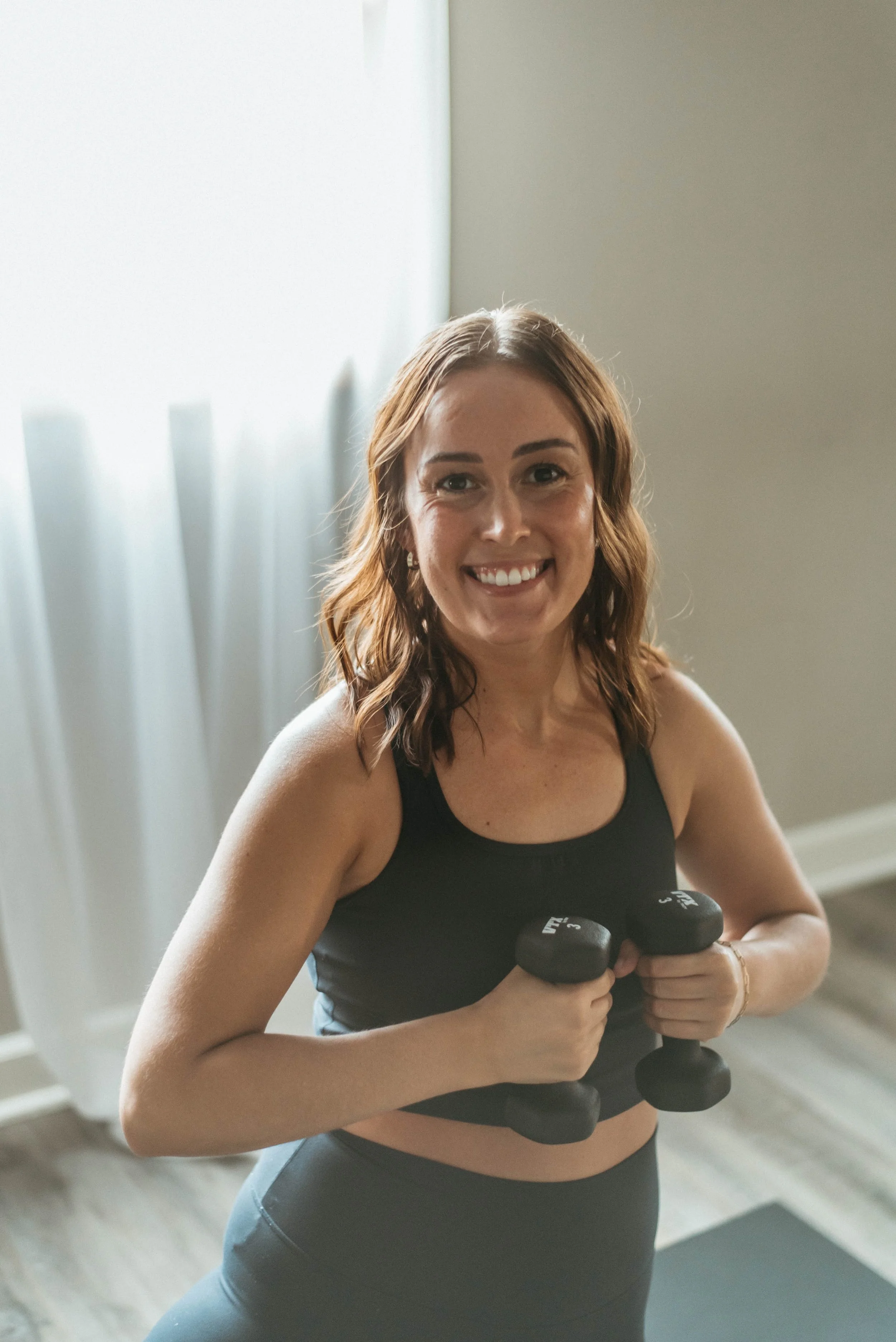 A woman sitting cross-legged on a yoga mat in a well-lit room, smiling at the camera.