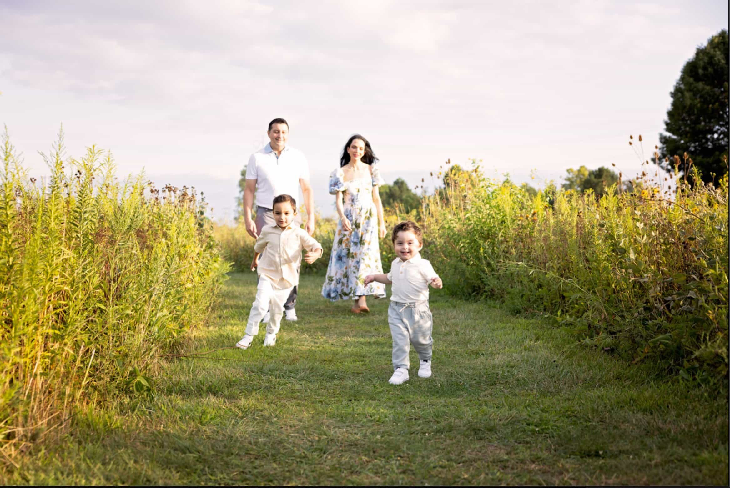 A family of four running and walking on a grassy path in a field of tall yellow and green plants, with two children in the front and two adults in the background, under a partly cloudy sky.