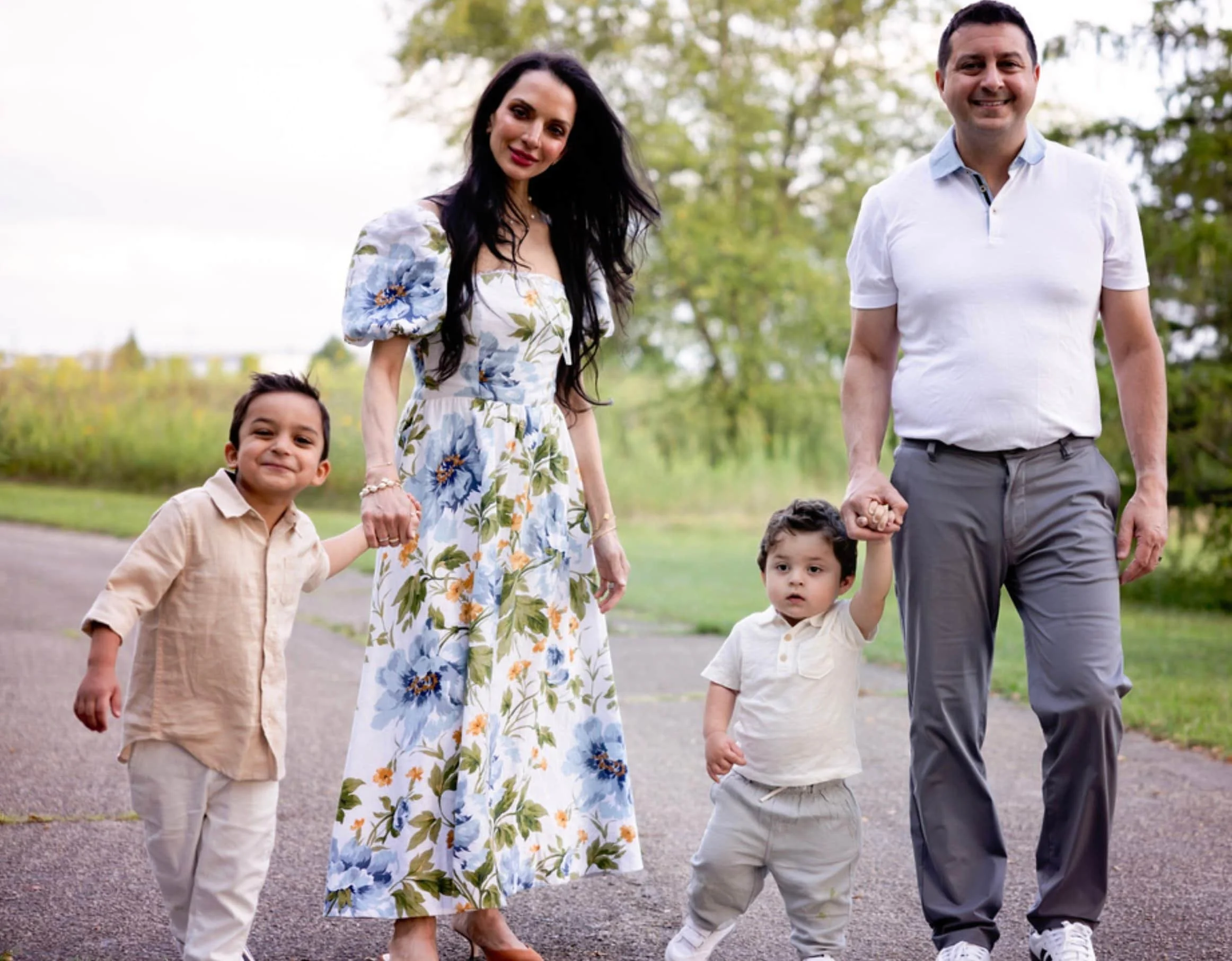 A family of four walking outdoors on a path in a park, holding hands. The mother is wearing a long white dress with blue floral patterns, and the father is dressed in a white polo shirt and gray pants. Two young boys, one in a beige shirt and the other in a white shirt, are holding their parents' hands.