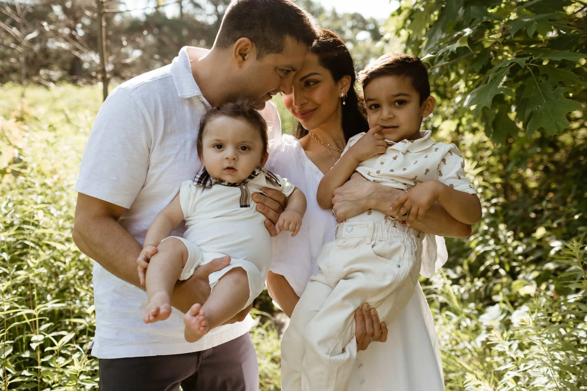 Family of four, two adults and two children, standing outdoors in a leafy green area during daytime. The adults are holding the children and leaning their heads close together, all smiling or looking content.