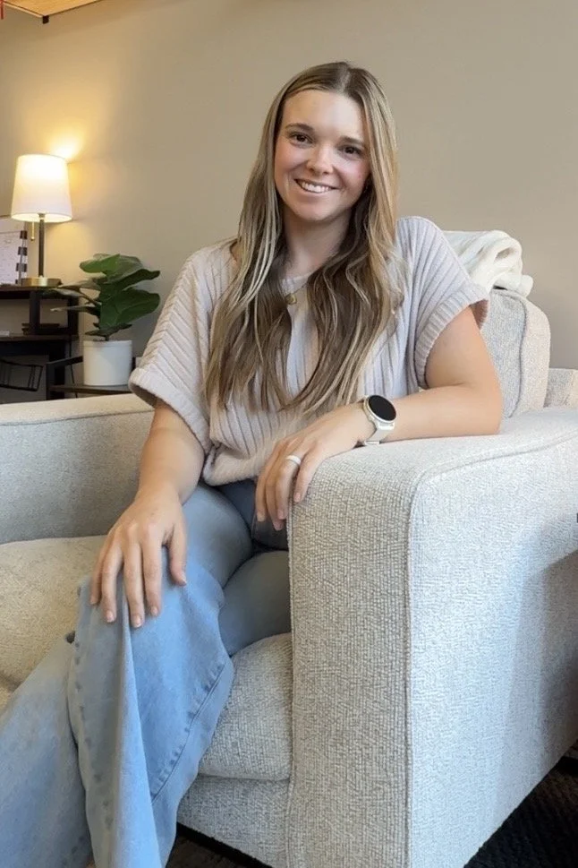 A young woman with long, wavy blonde hair sitting on a beige armchair in a cozy living room, smiling at the camera. She is wearing a light-colored, short-sleeved top, light blue jeans, a smartwatch, and a ring on her left hand. In the background, there is a side table with a lamp and a potted plant.