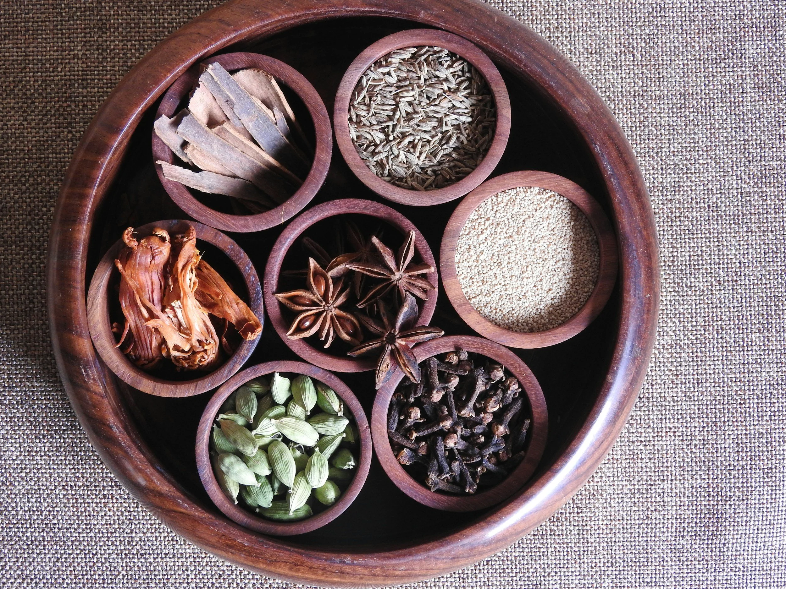A round wooden spice box containing various spices including cinnamon sticks, cumin seeds, star anise, coriander seeds, cloves, cardamom pods, fennel seeds, and another spice, on a textured surface.