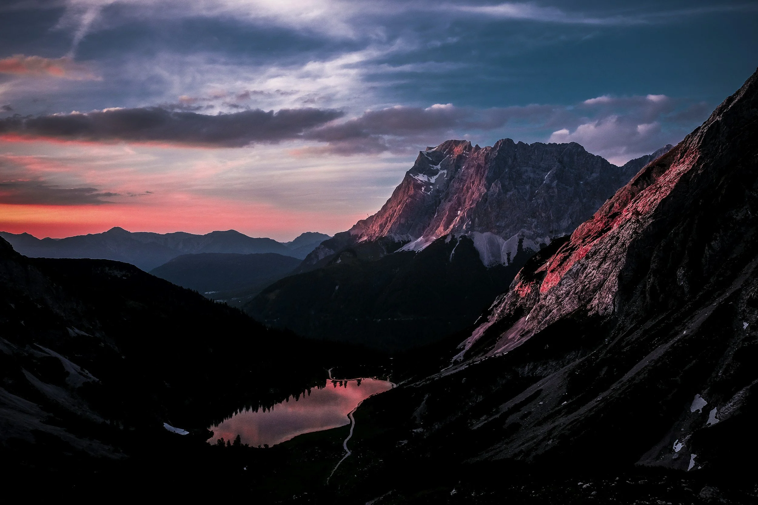 A mountain landscape at sunset with a pink and purple sky, rugged mountains with patches of snow, and a small reflective lake in the foreground.