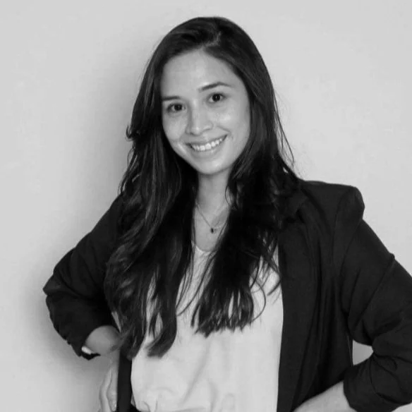 Black and white photo of a young woman with long wavy hair, smiling, wearing a light top and a dark jacket with her hands on her hips against a plain background.