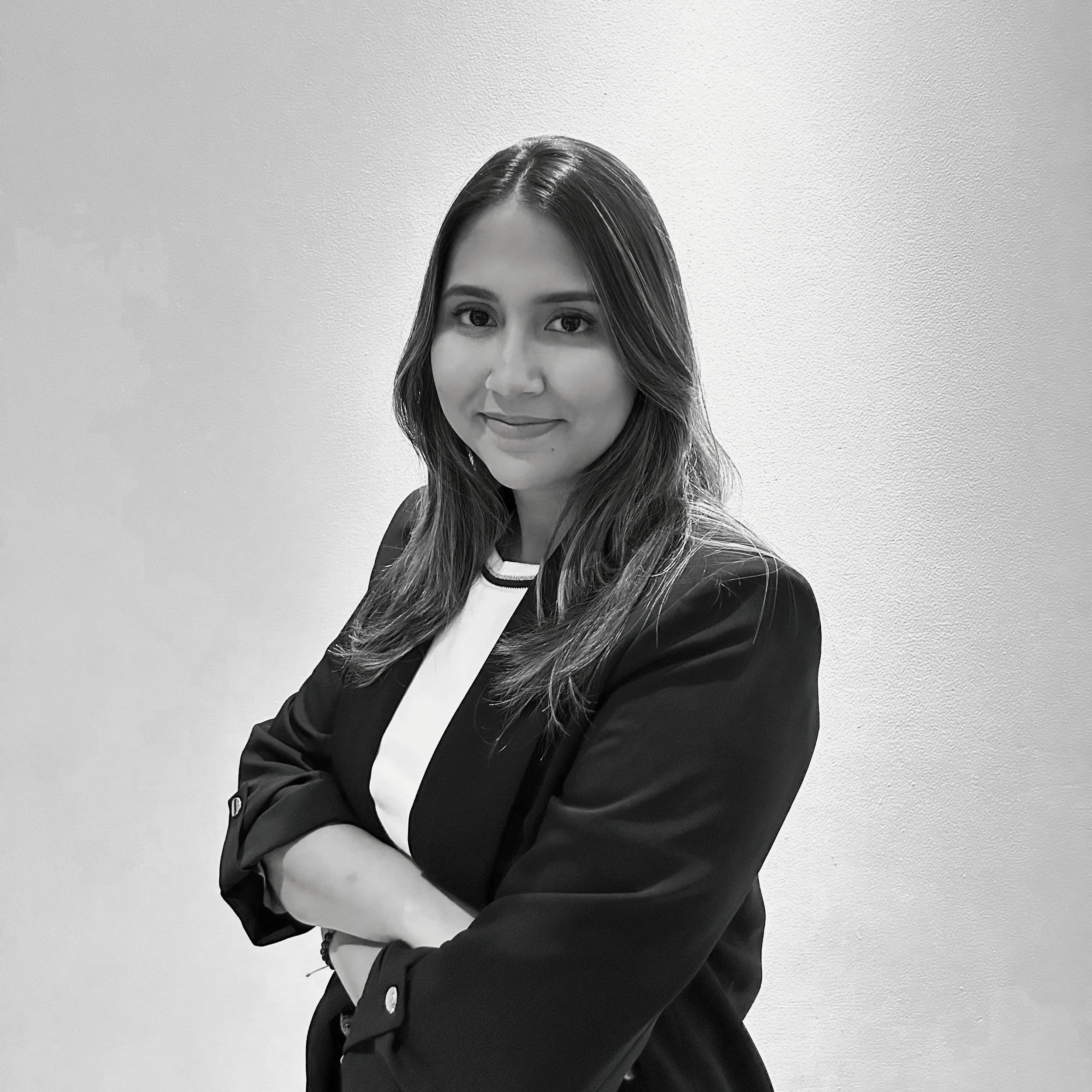 Black and white portrait of a woman with shoulder-length hair, arms crossed, smiling slightly, wearing a blazer over a shirt, standing against a plain wall.