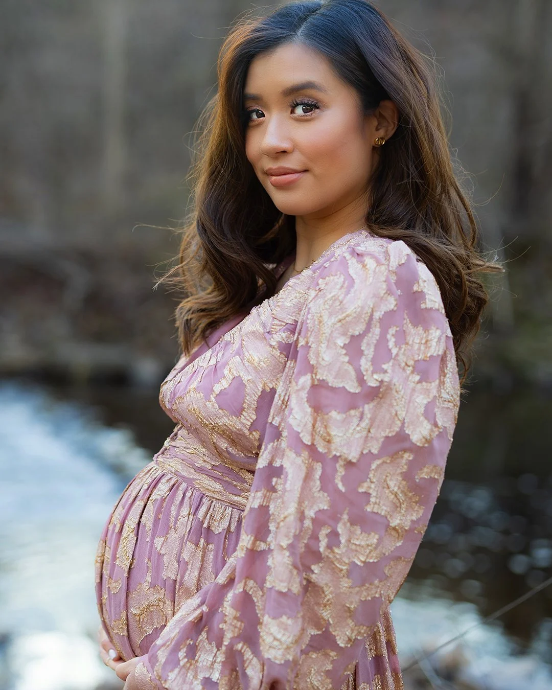 A pregnant woman wearing a pink and gold embroidered dress outdoors near a stream or river, standing with her hand on her belly and smiling softly.