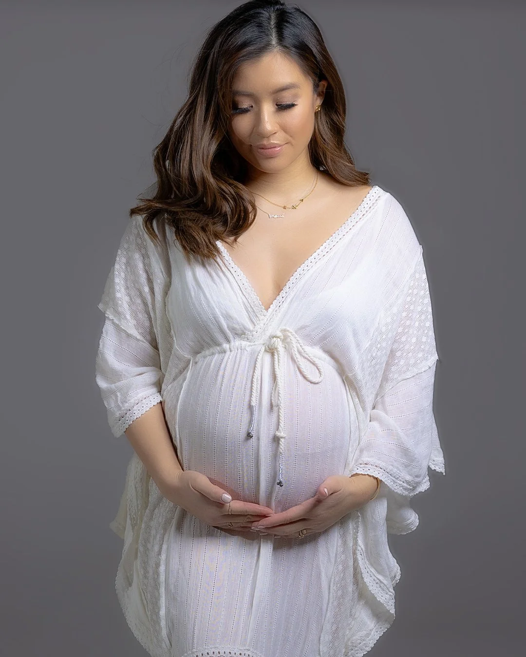A pregnant woman with long, wavy brown hair, wearing a white textured dress, gently holding her baby bump, standing against a neutral gray background.
