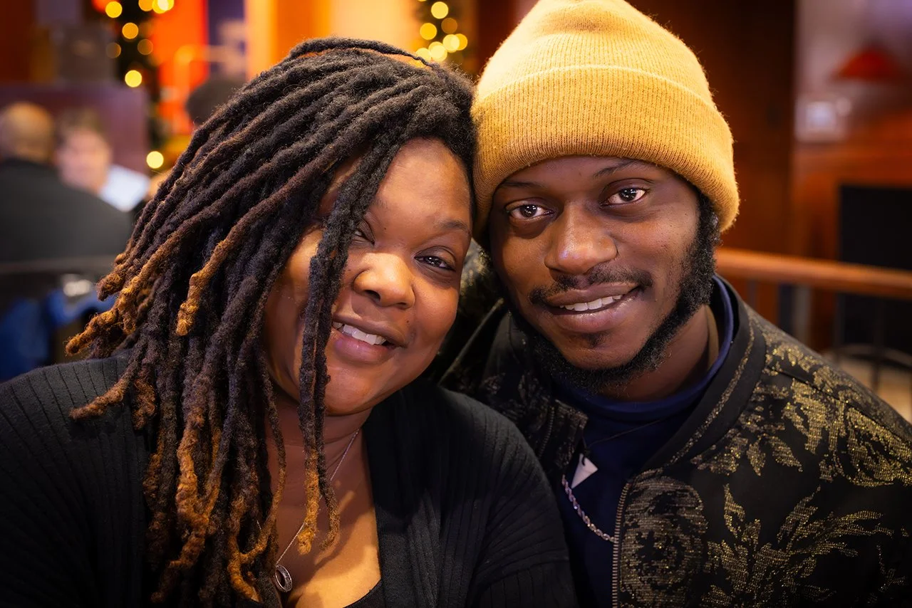 A woman and a man posing for a photography in a warmly lit indoor setting, with festive lights in the background.