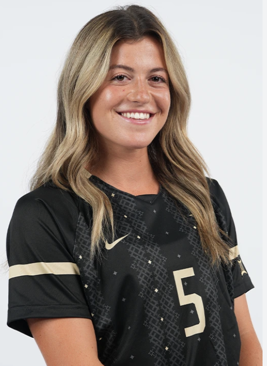 Young woman with long, wavy blond hair smiling, wearing a black sports jersey with gold and white accents, including the number 5, against a plain white background.