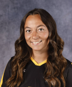 Portrait of a young woman with long wavy brown hair, wearing a black sports jersey with yellow trim, smiling against a gray background.