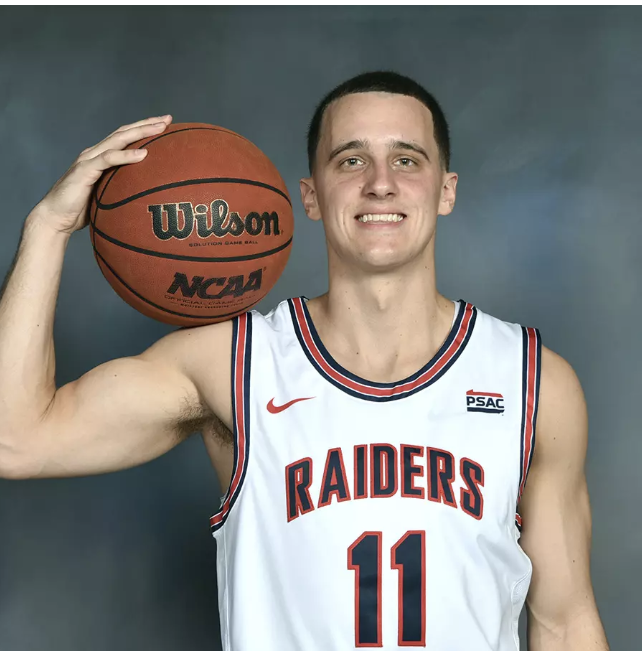 Young male basketball player holding a basketball on his shoulder, wearing a white Raiders jersey with the number 11.