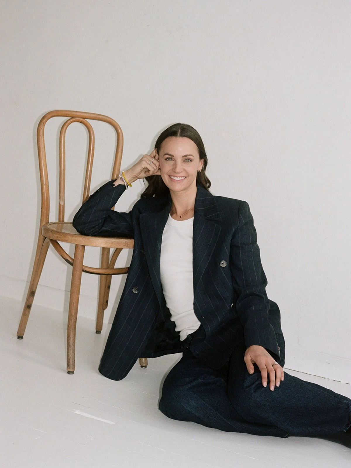 Woman sits playfully on the studio floor beside a bentwood chair in a pinstripe suit