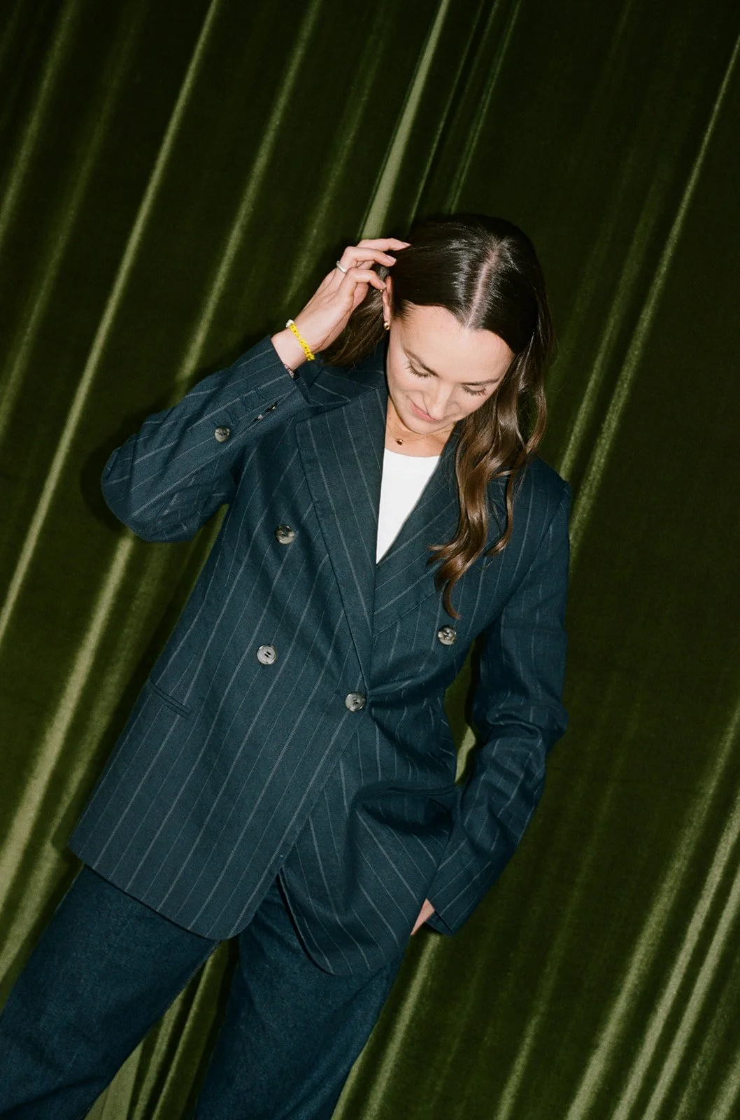 Woman in a pinstripe blazer runs a hand through her hair against a dark green velvet curtain