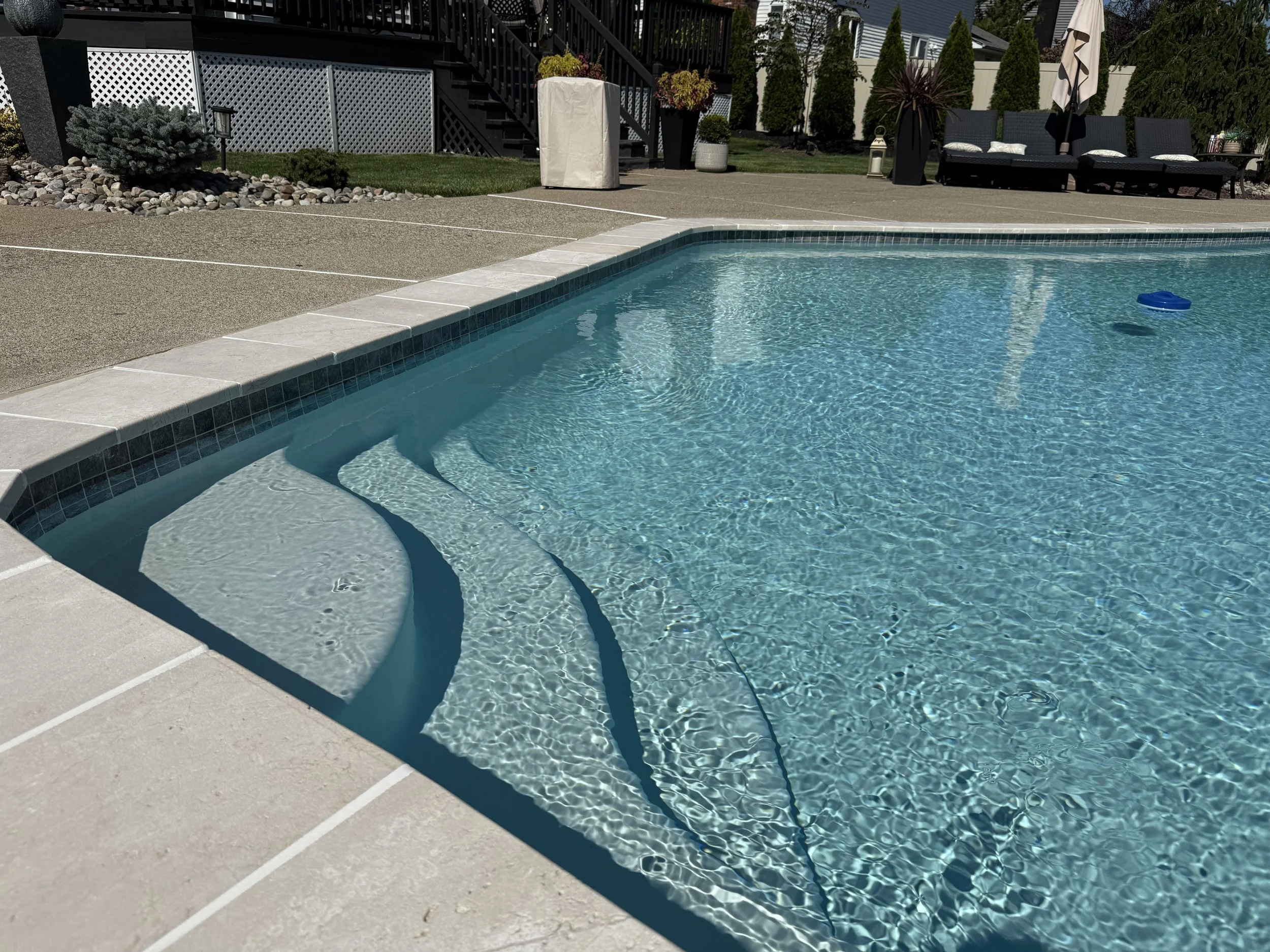A backyard swimming pool with clear water, a small set of water steps at the corner, surrounded by concrete and tile decking. In the background, there are outdoor lounge chairs, potted plants, and a patio with umbrellas and grass.