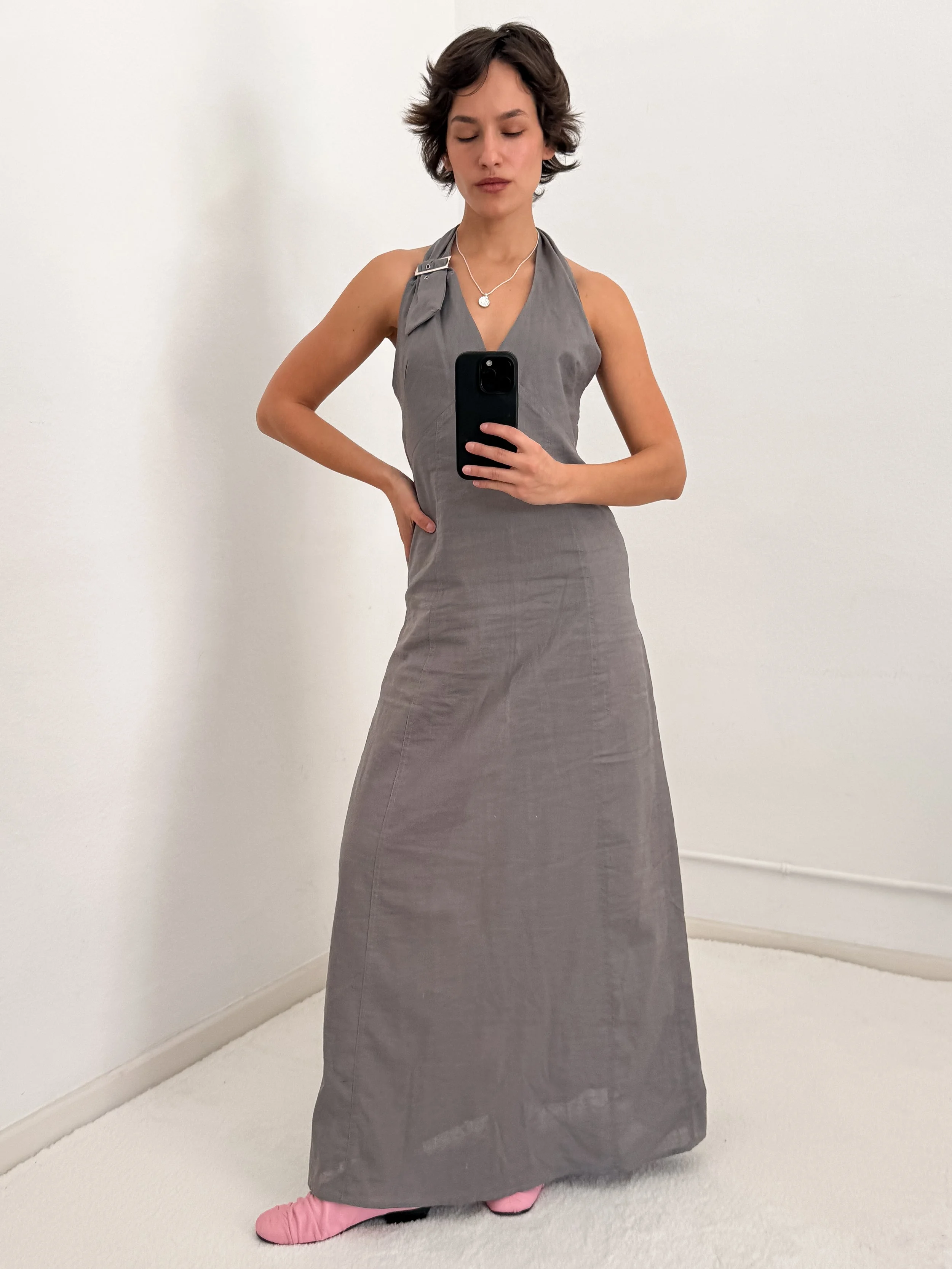 A woman taking a mirror selfie in a plain white room, wearing a sleeveless gray dress and pink slippers.