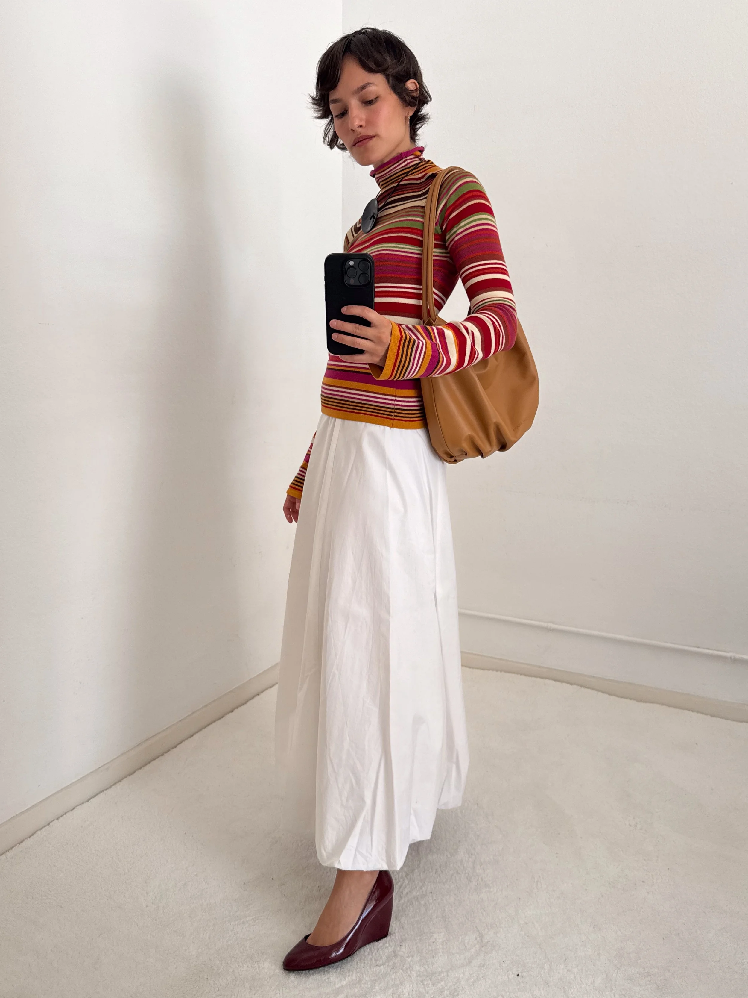A woman takes a mirror selfie against a plain white wall, wearing a colorful striped turtleneck, long white skirt, maroon wedge heels, a brown bag, and a round black accessory on her chest.