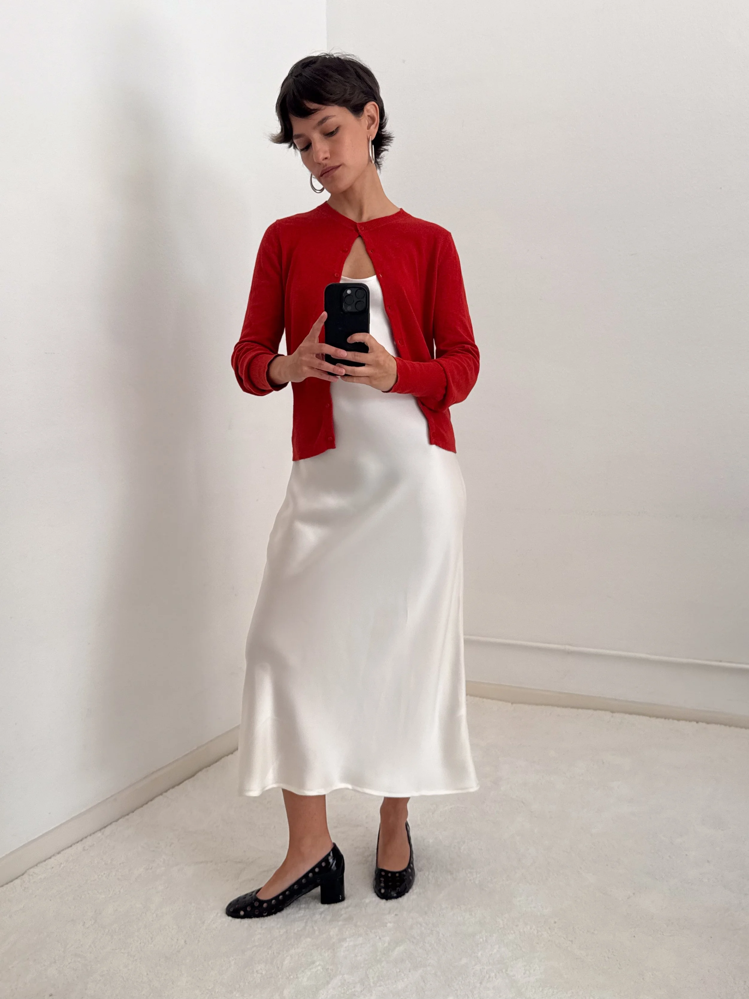 Woman wearing black high heels with open toe and polka dot pattern, white satin midi skirt, white top, red cardigan, silver hoop earrings, holding a smartphone, standing in a white room.
