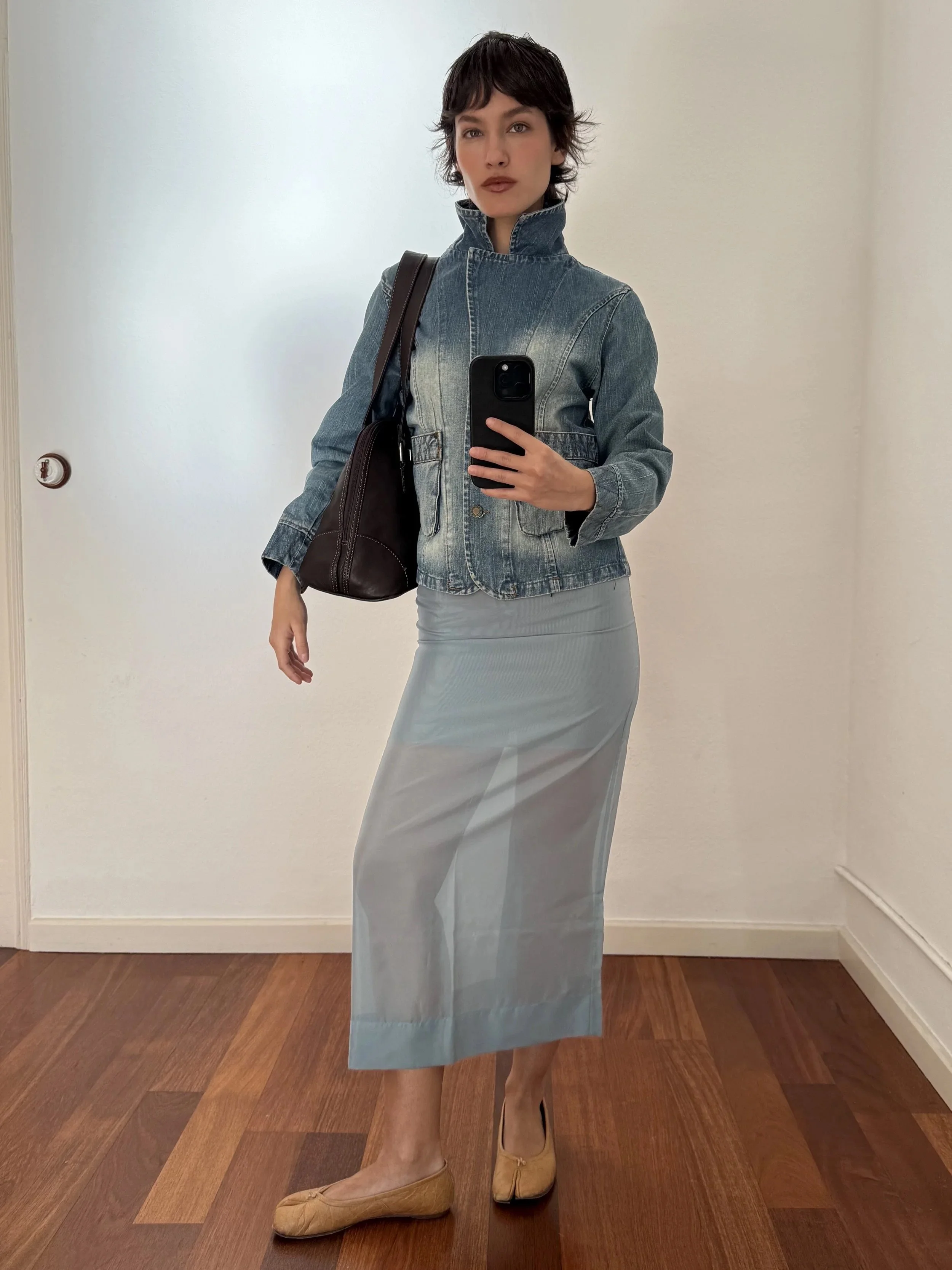 A woman taking a mirror selfie wearing a denim jacket, gray skirt, and beige shoes, holding a dark bag over her shoulder in a room with hardwood floors and plain white walls.