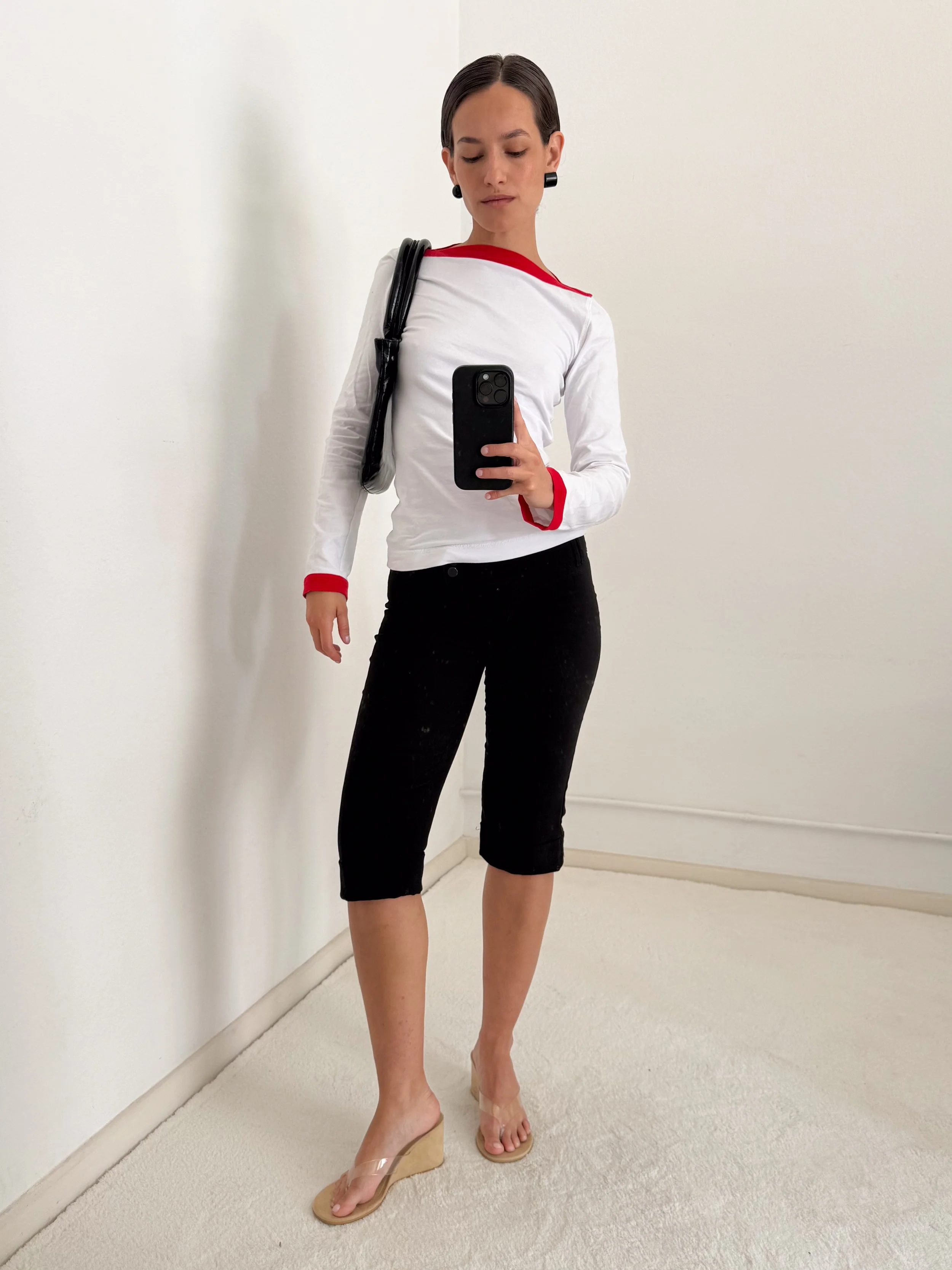 A woman taking a mirror selfie in an all-white room. She is wearing a white long-sleeve shirt with red accents, black pants, and clear high-heeled shoes. She is carrying a black shoulder bag and has short, dark hair.