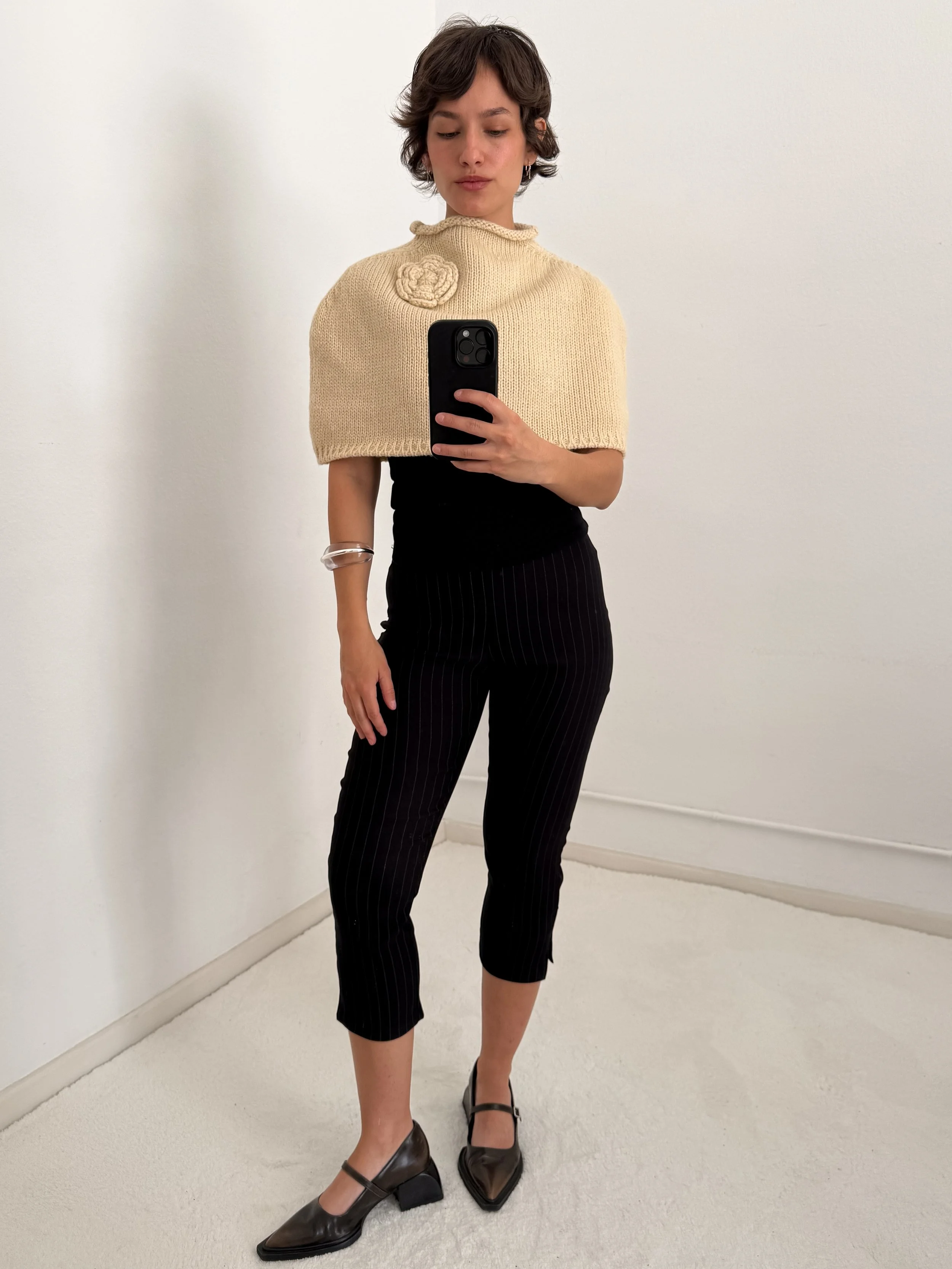A woman taking a mirror selfie wearing a beige knitted cape with a flower decoration on the shoulder, black pinstripe pants, and brown high-heeled shoes.