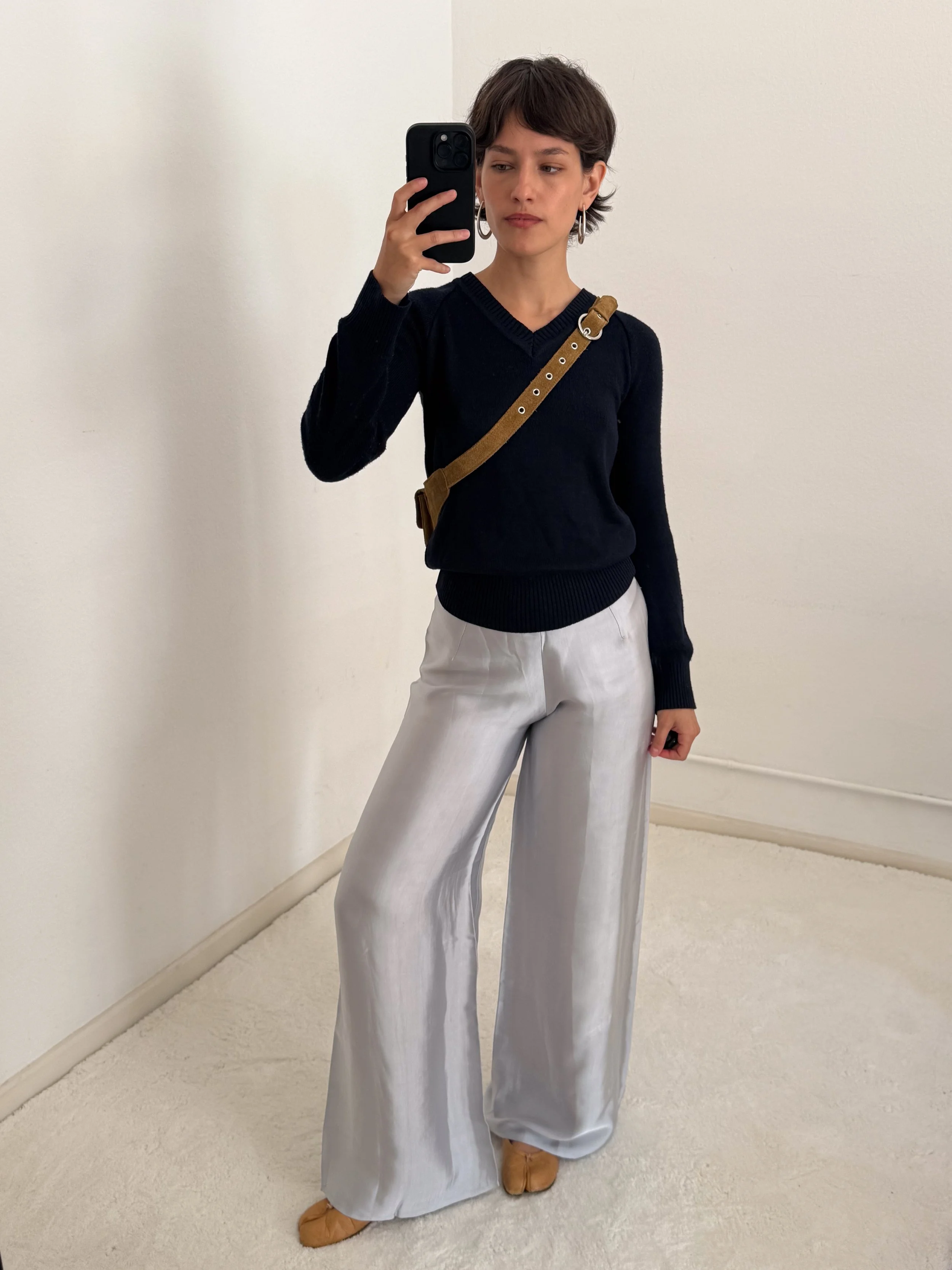 A woman taking a mirror selfie against a plain white wall, wearing a black V-neck sweater, white wide-leg pants, tan shoes, large hoop earrings, and carrying a brown shoulder bag.