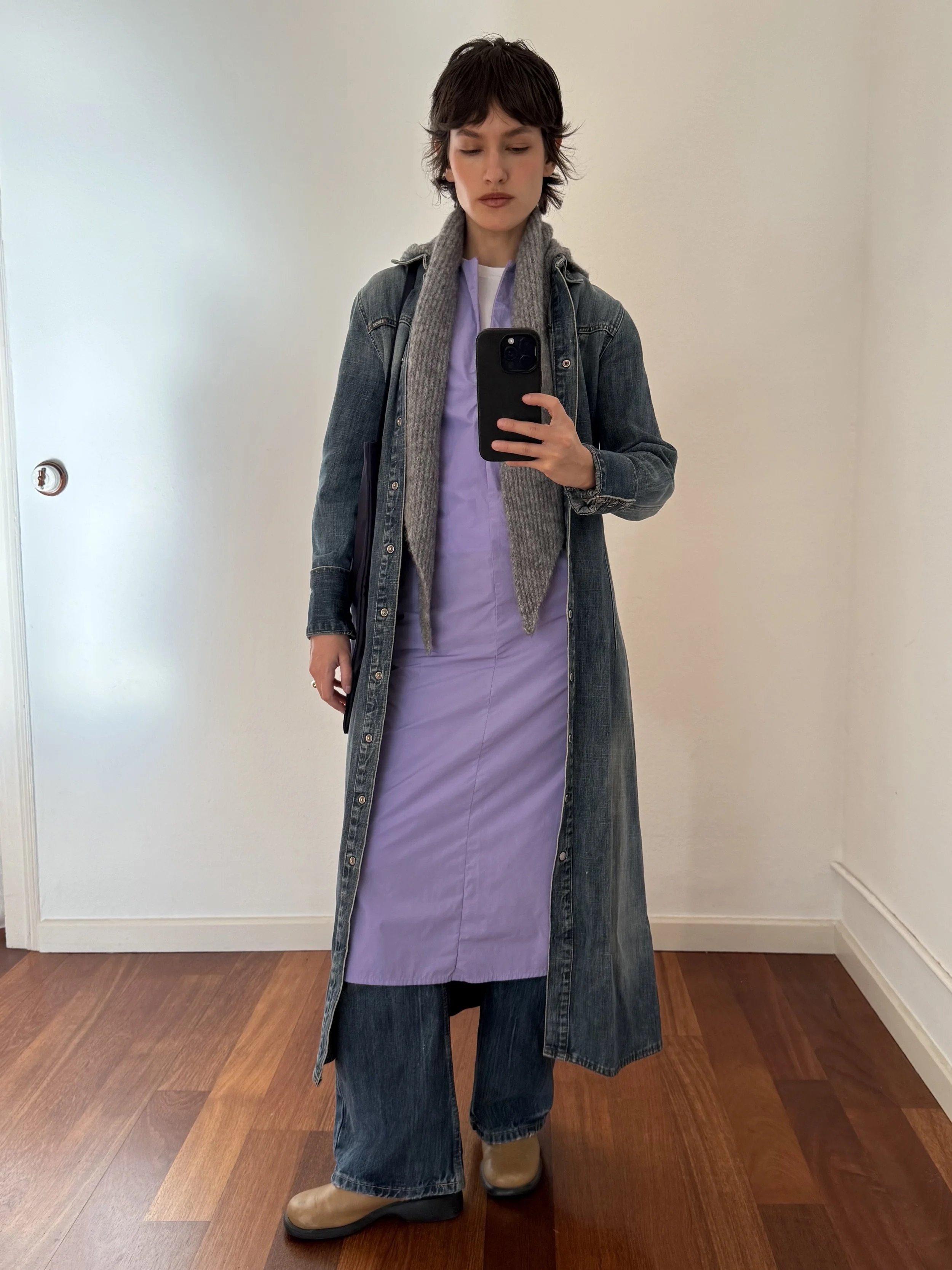 Person taking a mirror selfie wearing a denim jacket, purple dress, gray scarf, dark wide-leg jeans, and beige boots in a room with wooden flooring and plain white walls.