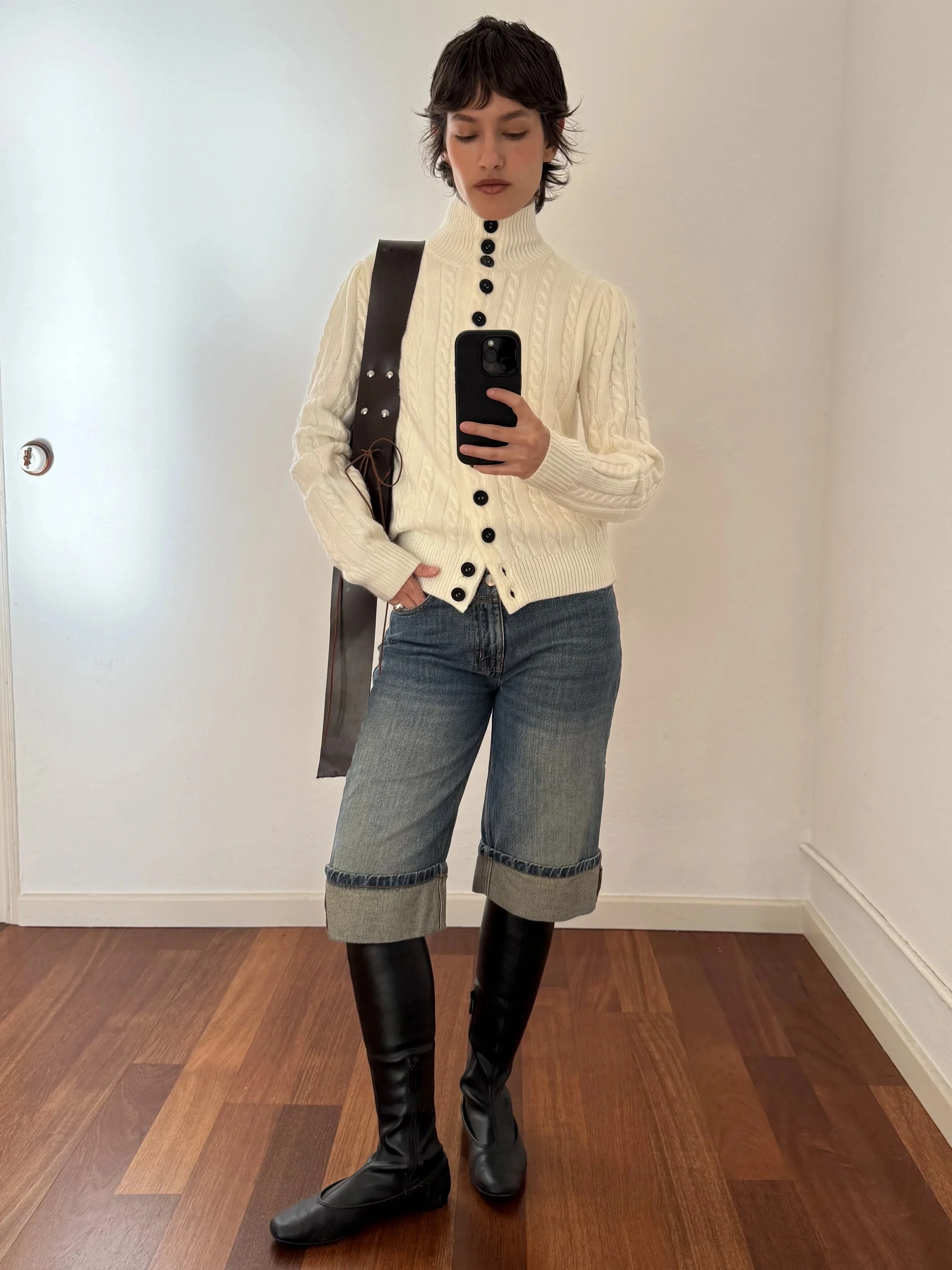 A person taking a mirror selfie wearing a cream-colored cable knit sweater with black buttons, blue cuffed jeans, and black knee-high boots, carrying a dark brown shoulder bag.