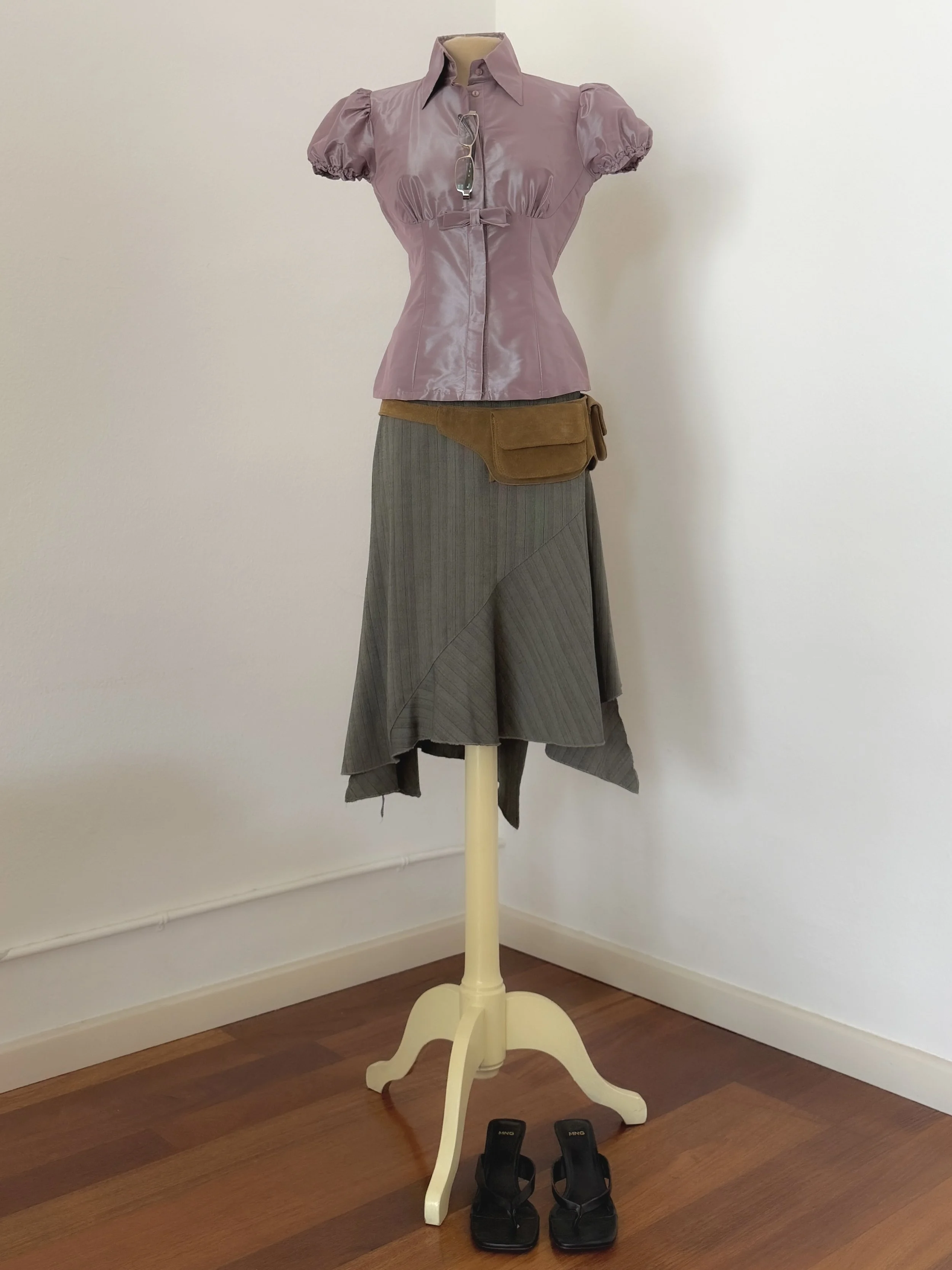 A mannequin dressed in a purple short-sleeve blouse, gray skirt, with black sandals positioned on the wooden floor in front of a white wall.