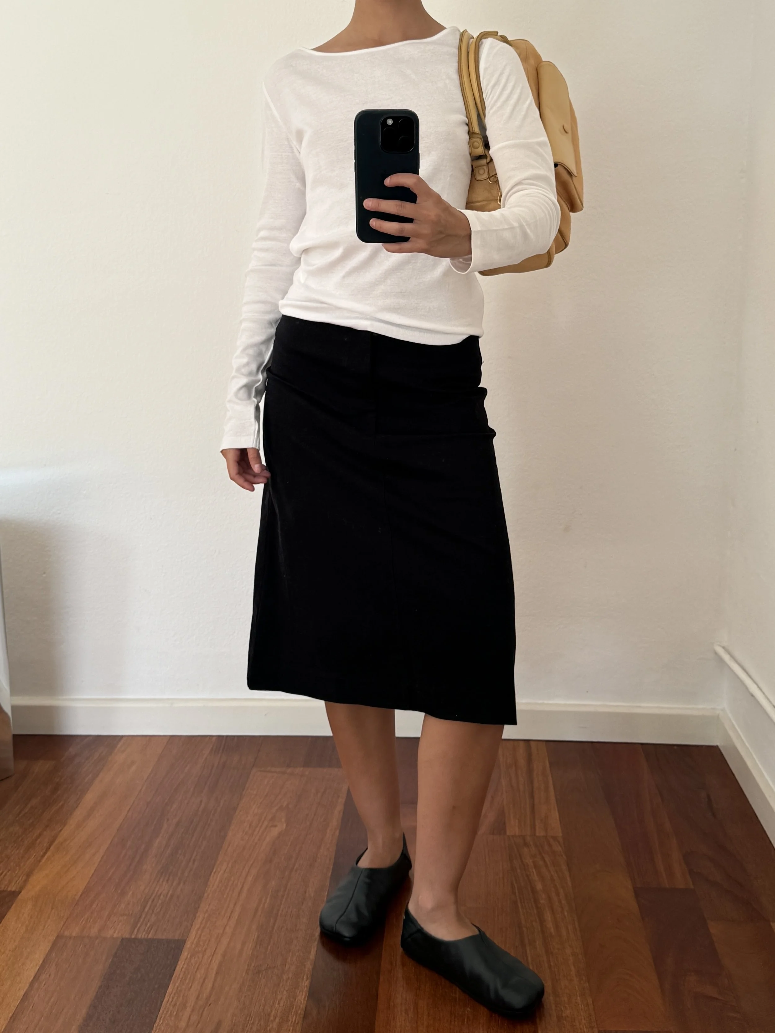 Person taking a mirror selfie wearing a white long-sleeve shirt, black skirt, black slippers, and carrying a tan backpack, with hardwood floor and plain white wall in the background.