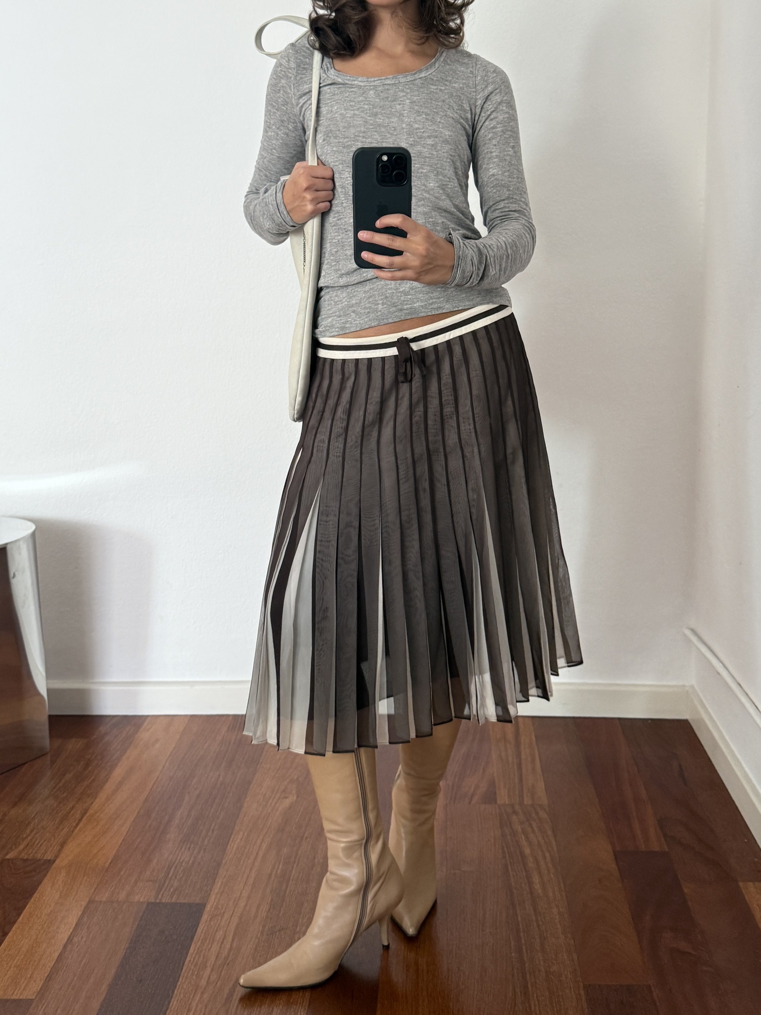A woman taking a mirror selfie wearing a gray long sleeve top, a black and white pleated midi skirt, and beige knee-high boots.