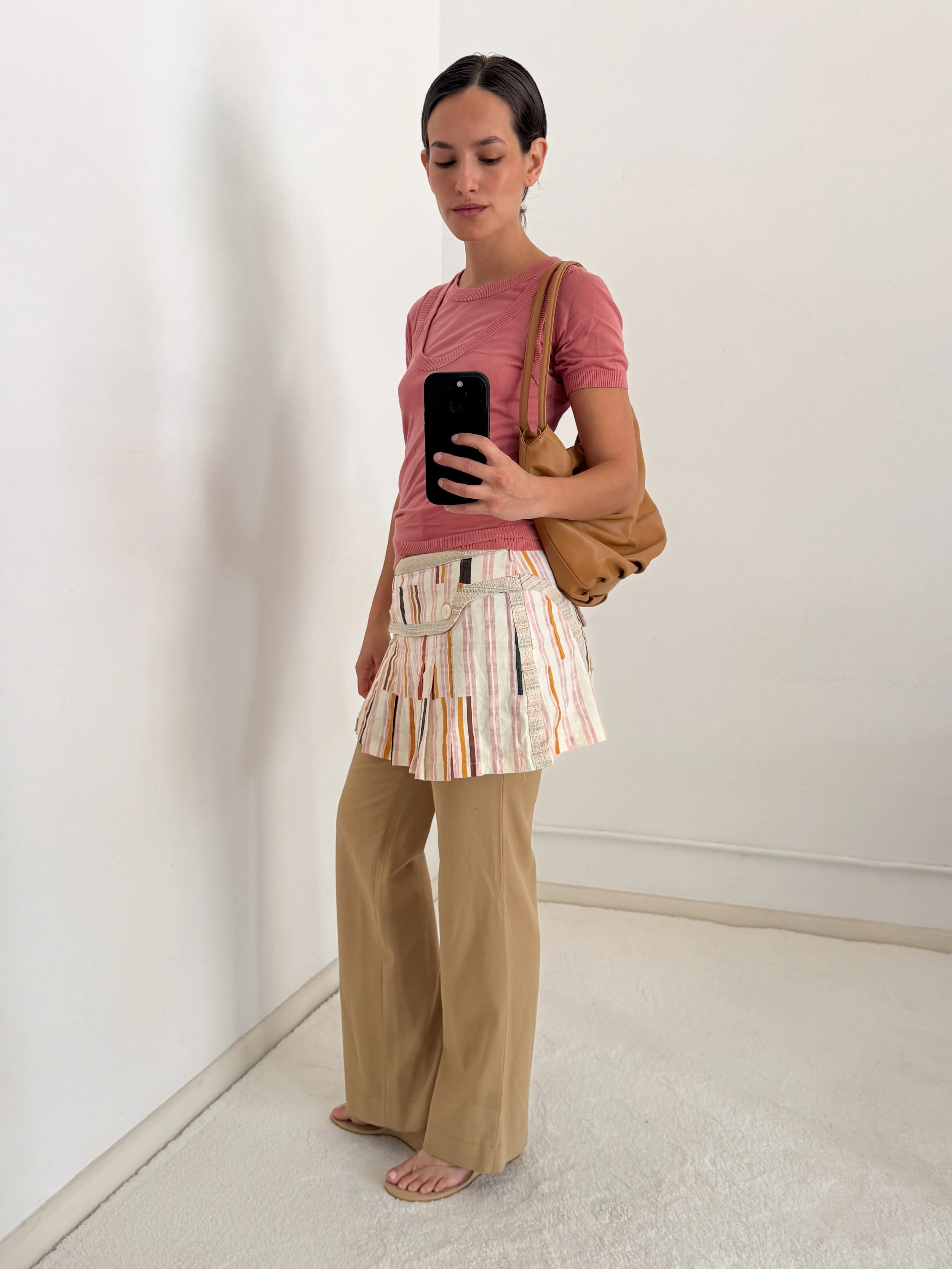A woman standing indoors taking a mirror selfie with her phone, wearing a pink t-shirt, beige wide-leg pants, carrying a brown shoulder bag, and barefoot.