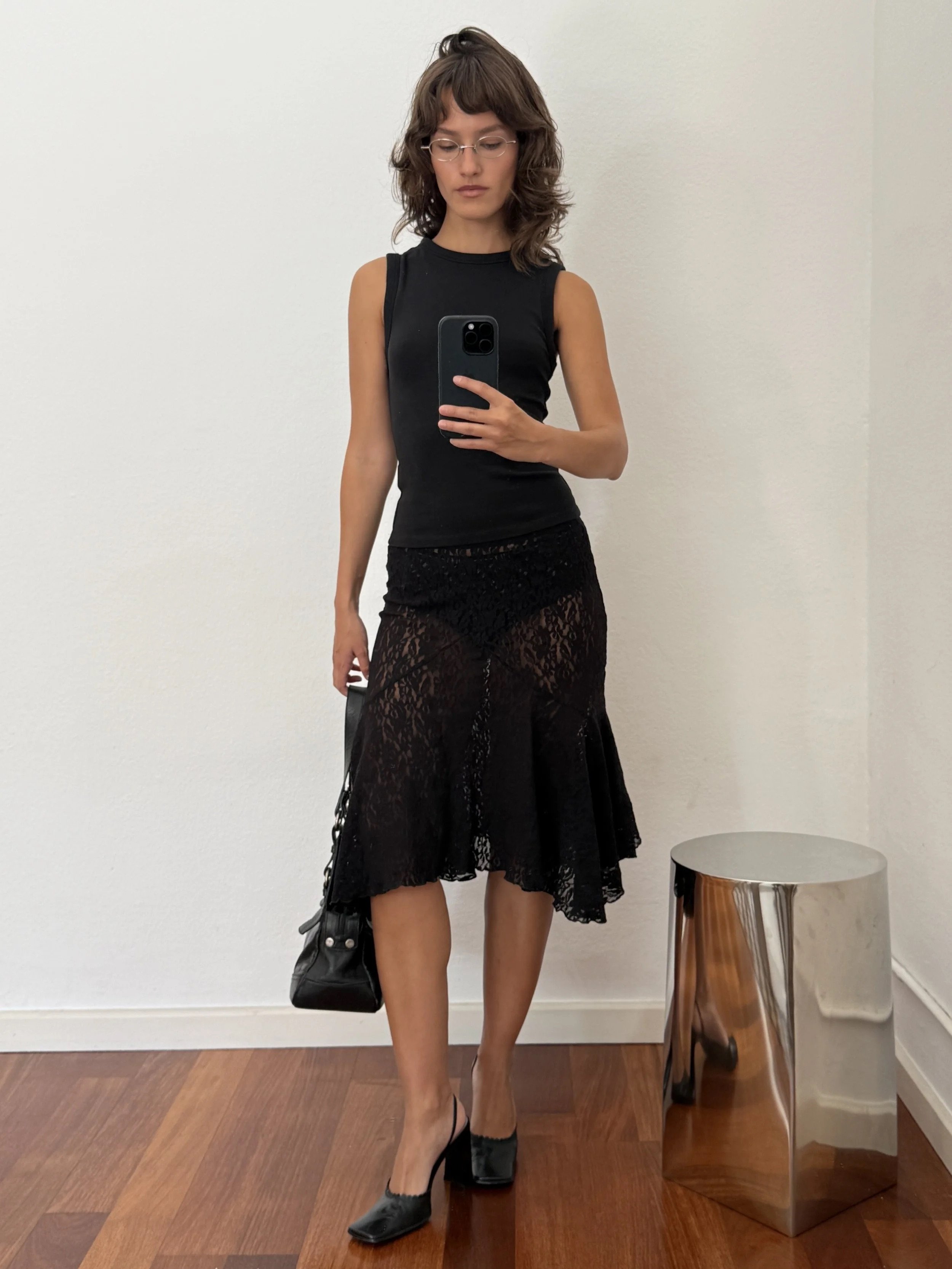 A young woman with brown hair and glasses taking a mirror selfie. She is wearing a black sleeveless top, a black lace skirt, and black shoes, and is carrying a black bag.