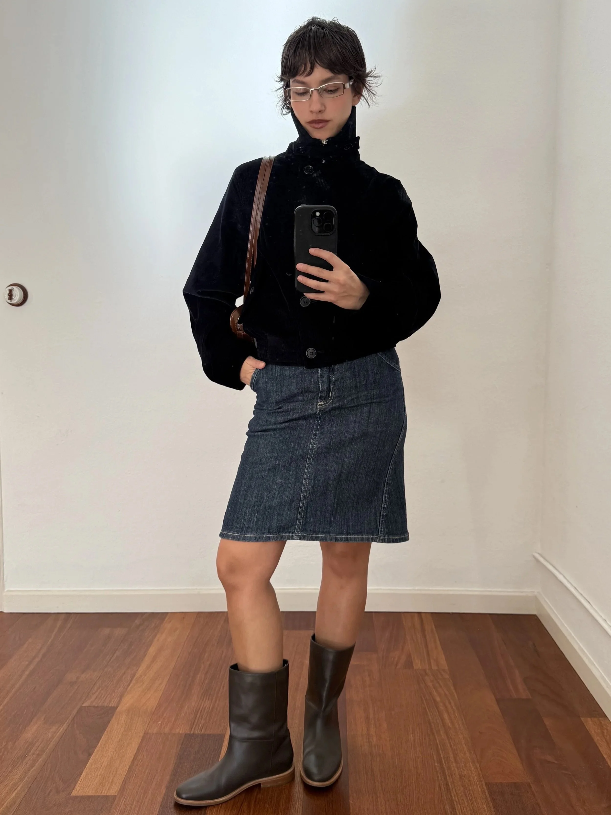 A woman with short wavy hair and glasses taking a mirror selfie. She is wearing a black velvet jacket, a denim skirt, and knee-high black boots. She has a brown shoulder bag.