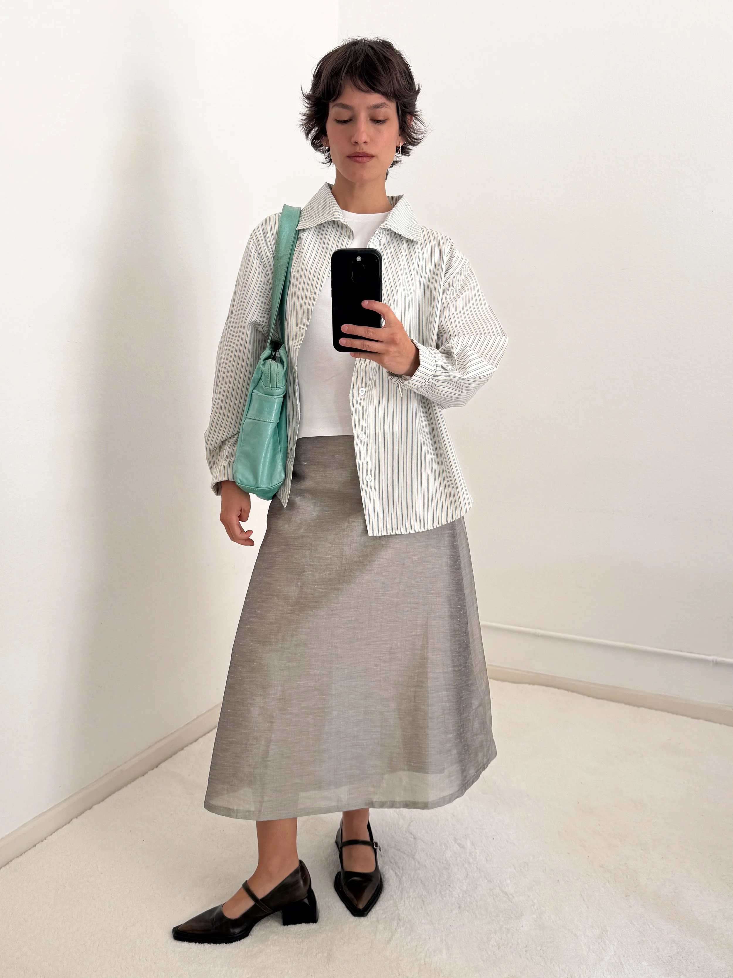 A woman taking a mirror selfie, wearing a striped button-up shirt over a white top, with a beige skirt, black pointed shoes, and carrying a teal bag, standing against a plain white wall.