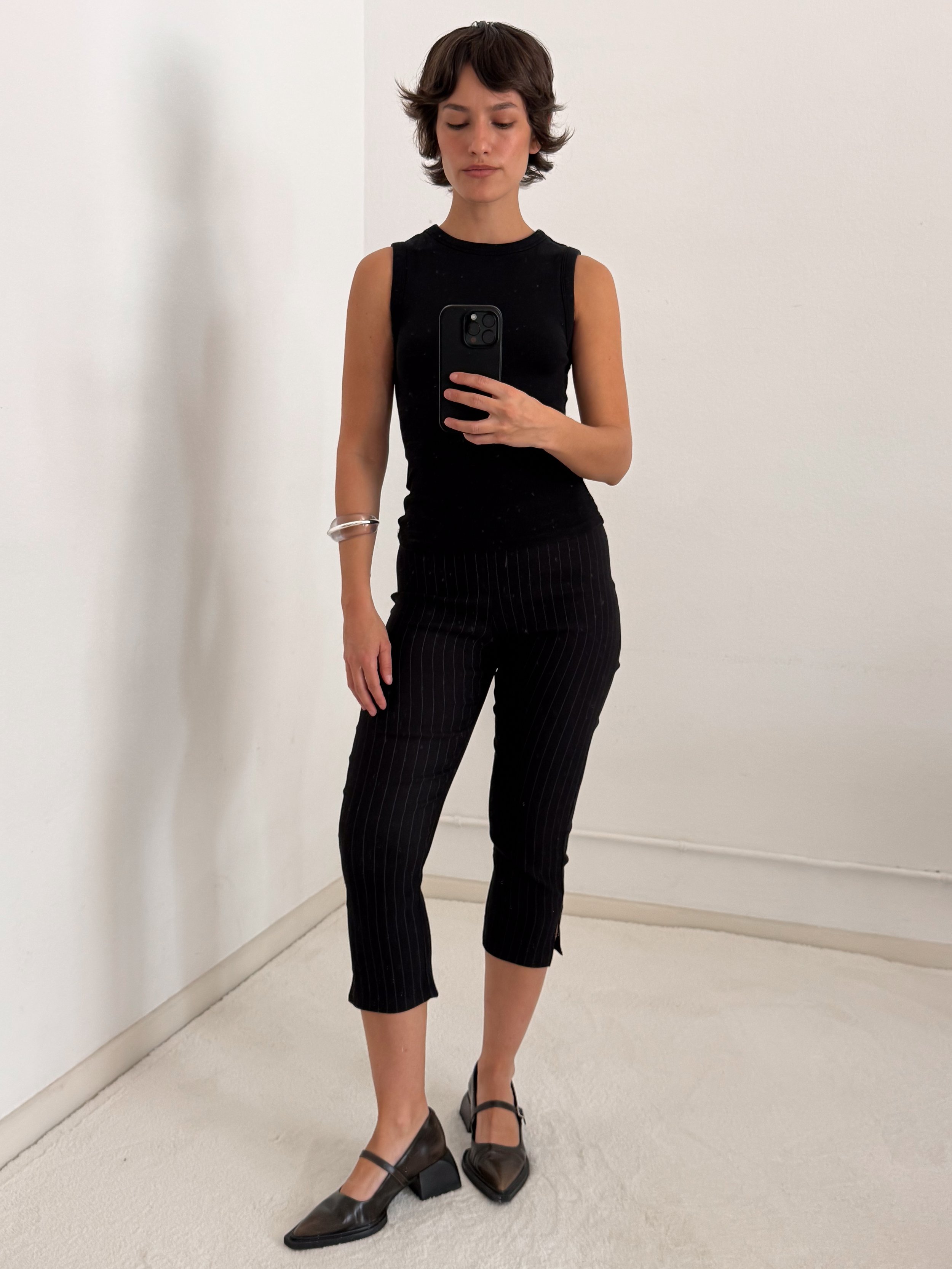 A woman standing in front of a white wall, wearing a sleeveless black top, pinstripe pants, and black block-heeled shoes, taking a mirror selfie with her phone.