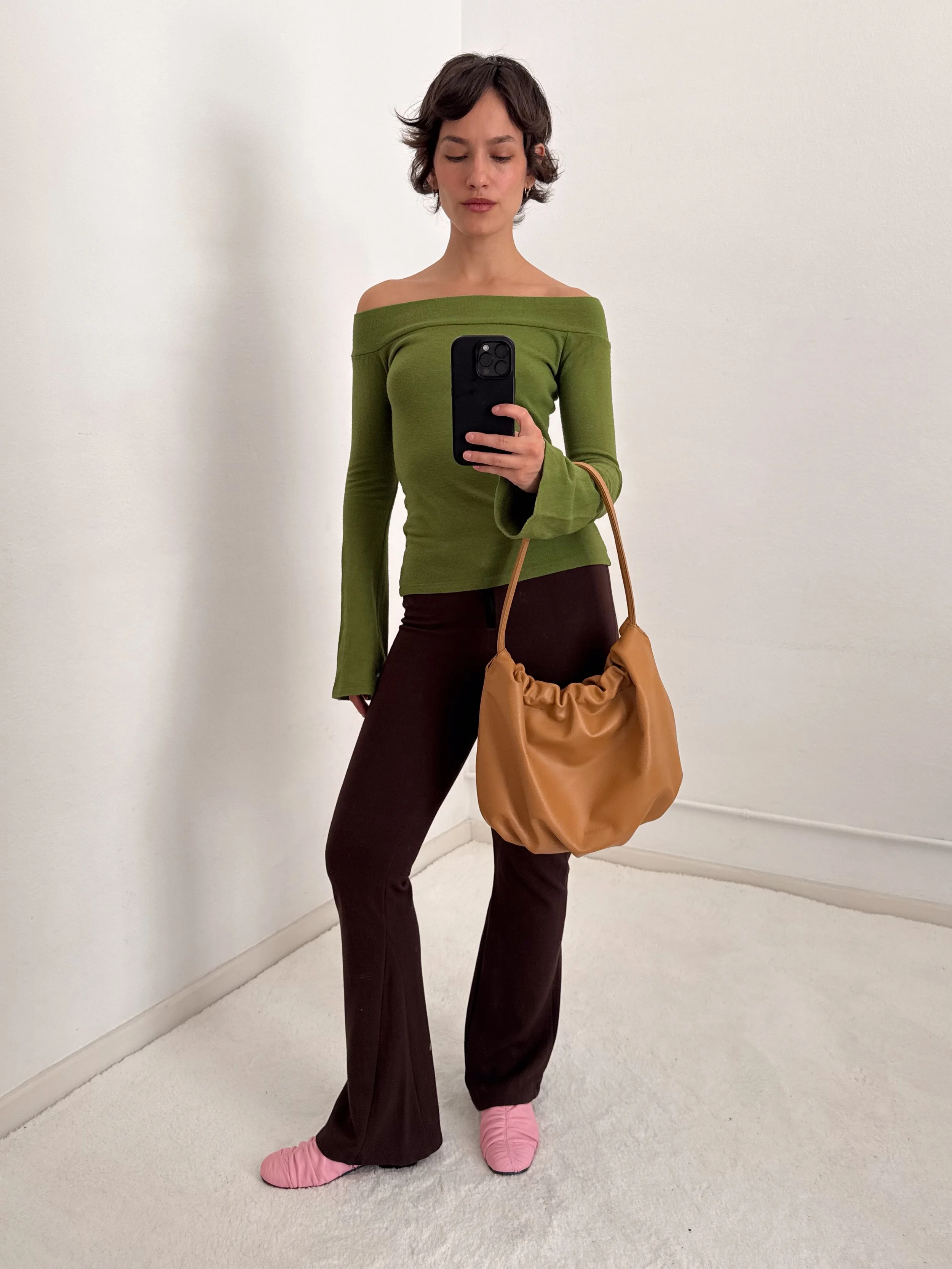 A woman taking a mirror selfie wearing a green off-the-shoulder top, dark brown pants, pink slippers, and holding a tan handbag against a plain white wall.