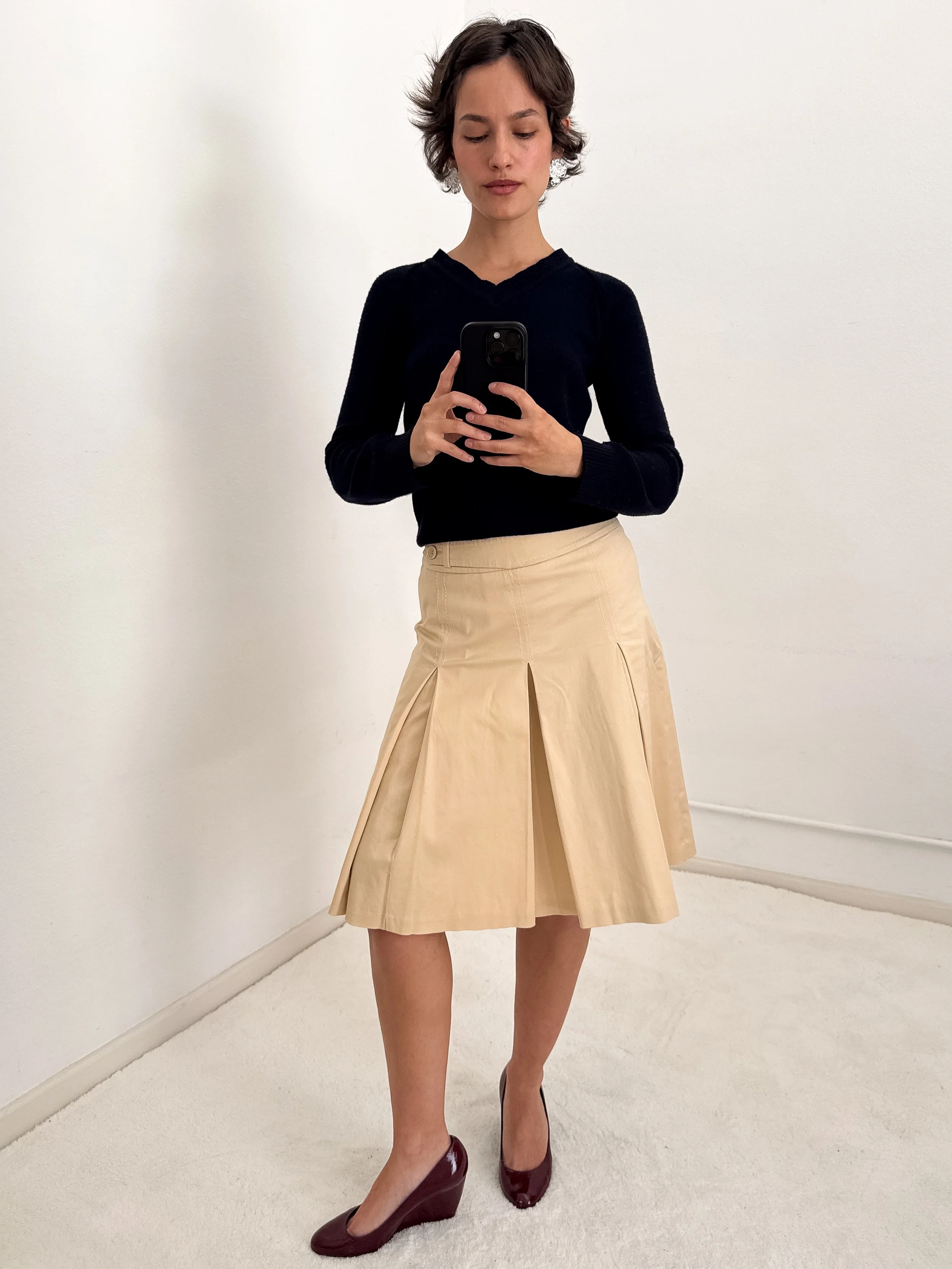 Woman taking a mirror selfie wearing a black top, beige pleated skirt, and maroon high heels, standing against a plain white wall.