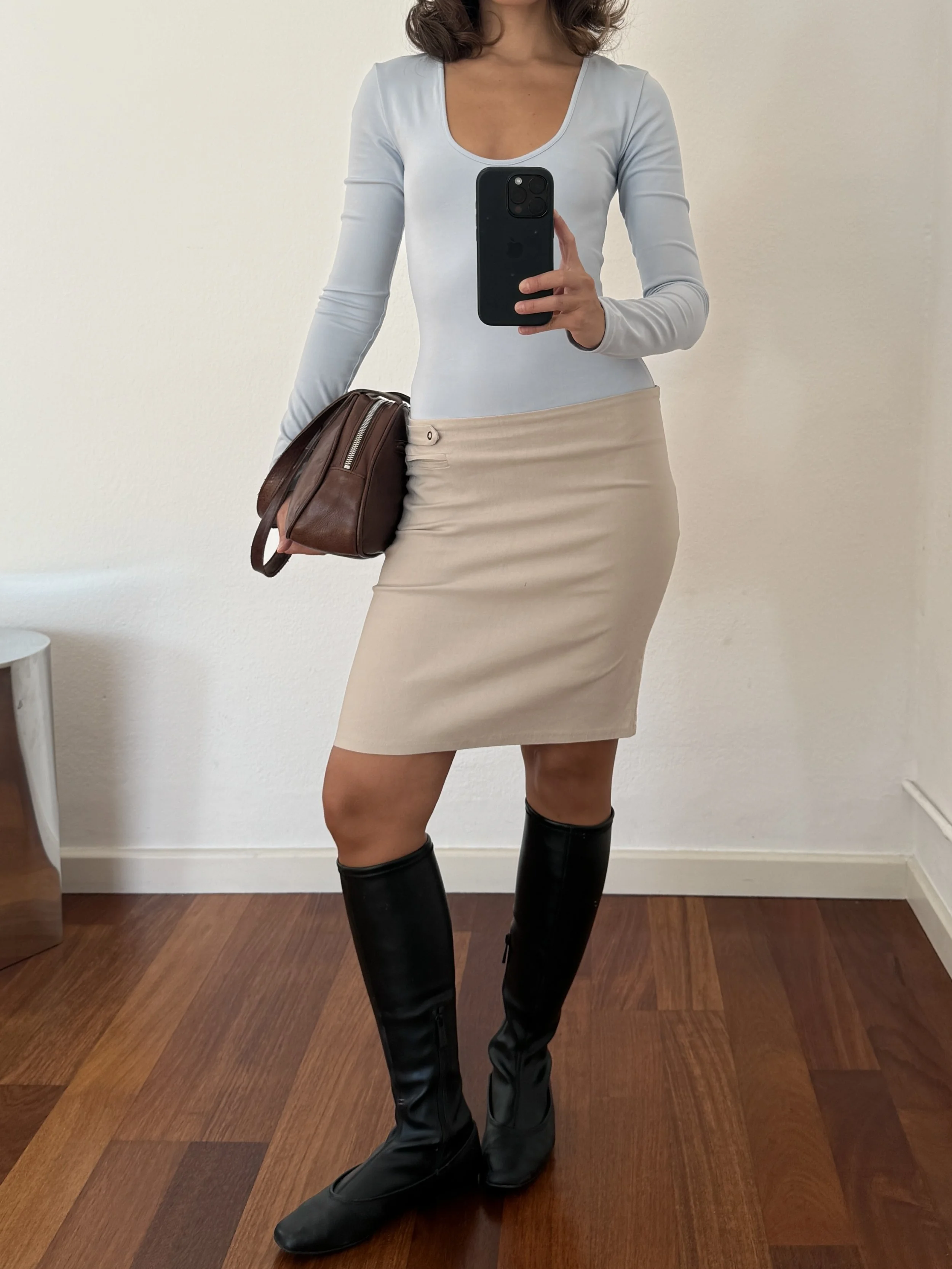 A woman taking a mirror selfie wearing a light blue long-sleeve shirt, beige skirt, black knee-high boots, and holding a brown handbag.