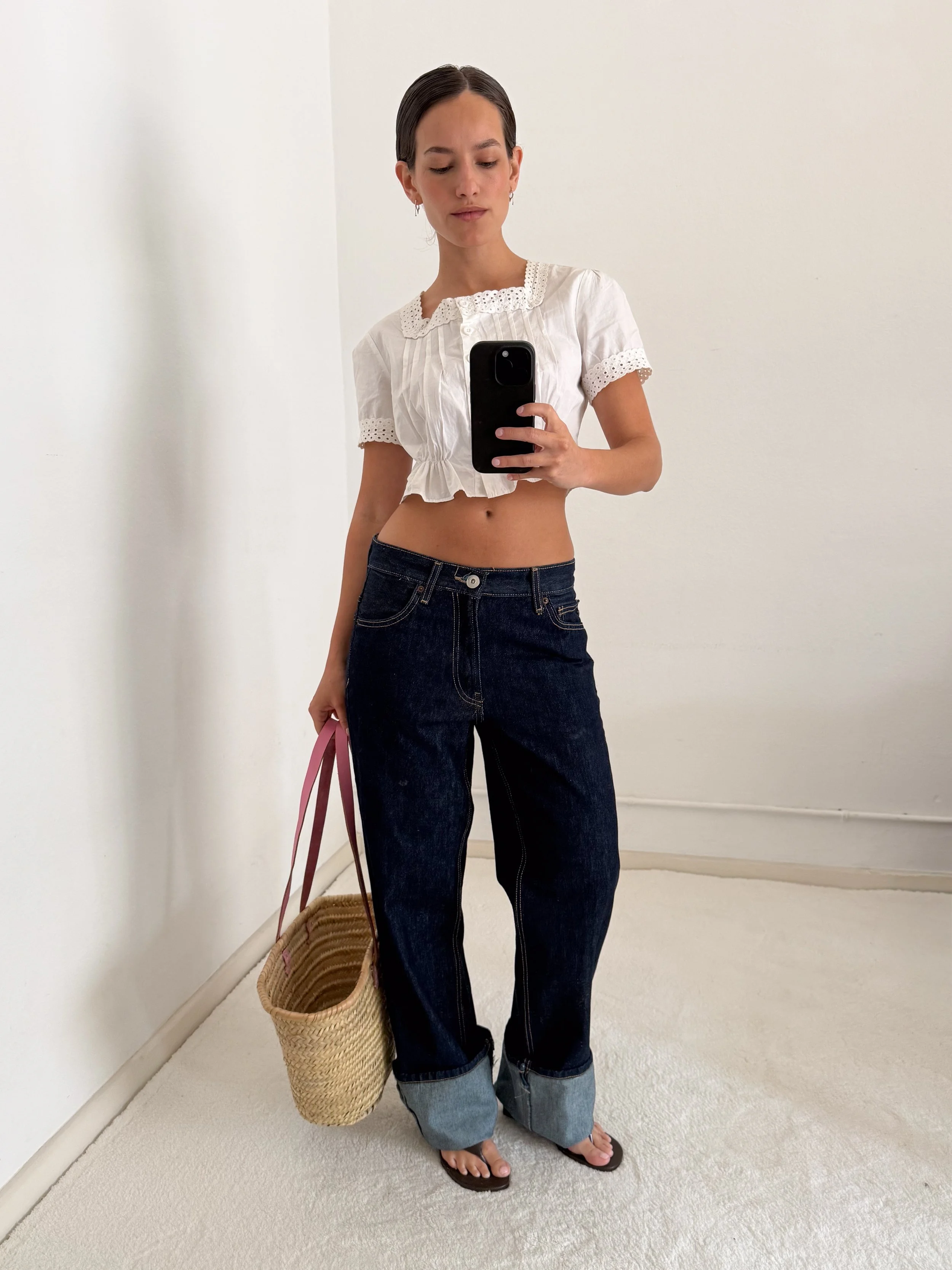 A woman standing on a white carpeted floor, taking a mirror selfie with her phone. She is wearing a white, short-sleeved, eyelet blouse, dark high-waisted jeans with rolled cuffs, black flip-flops, and has a wicker bag on the floor beside her.