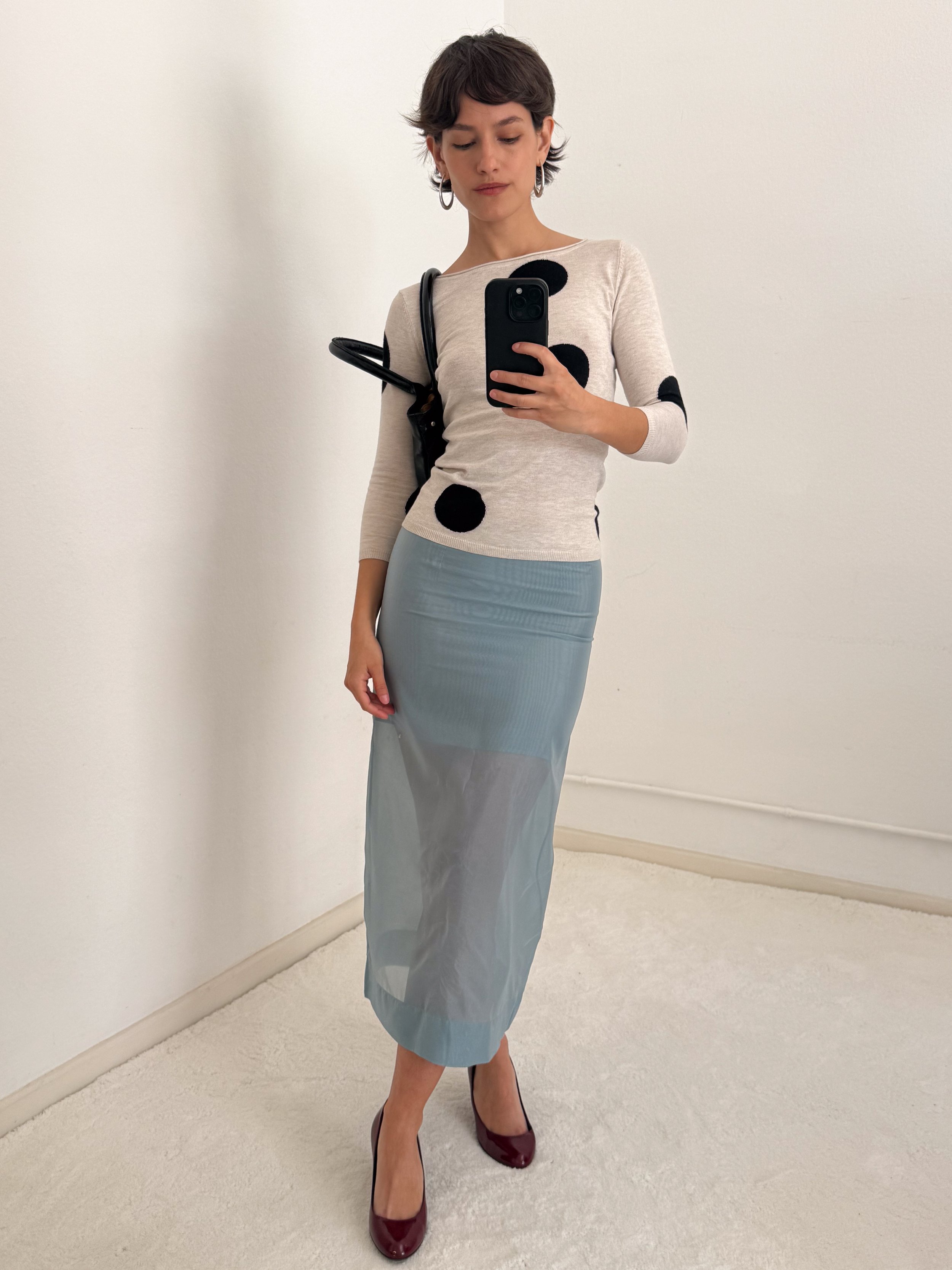A woman with short dark hair taking a selfie in a mirror, wearing a beige top with black polka dots, a light blue skirt, maroon high heels, and silver hoop earrings, against a plain white wall.