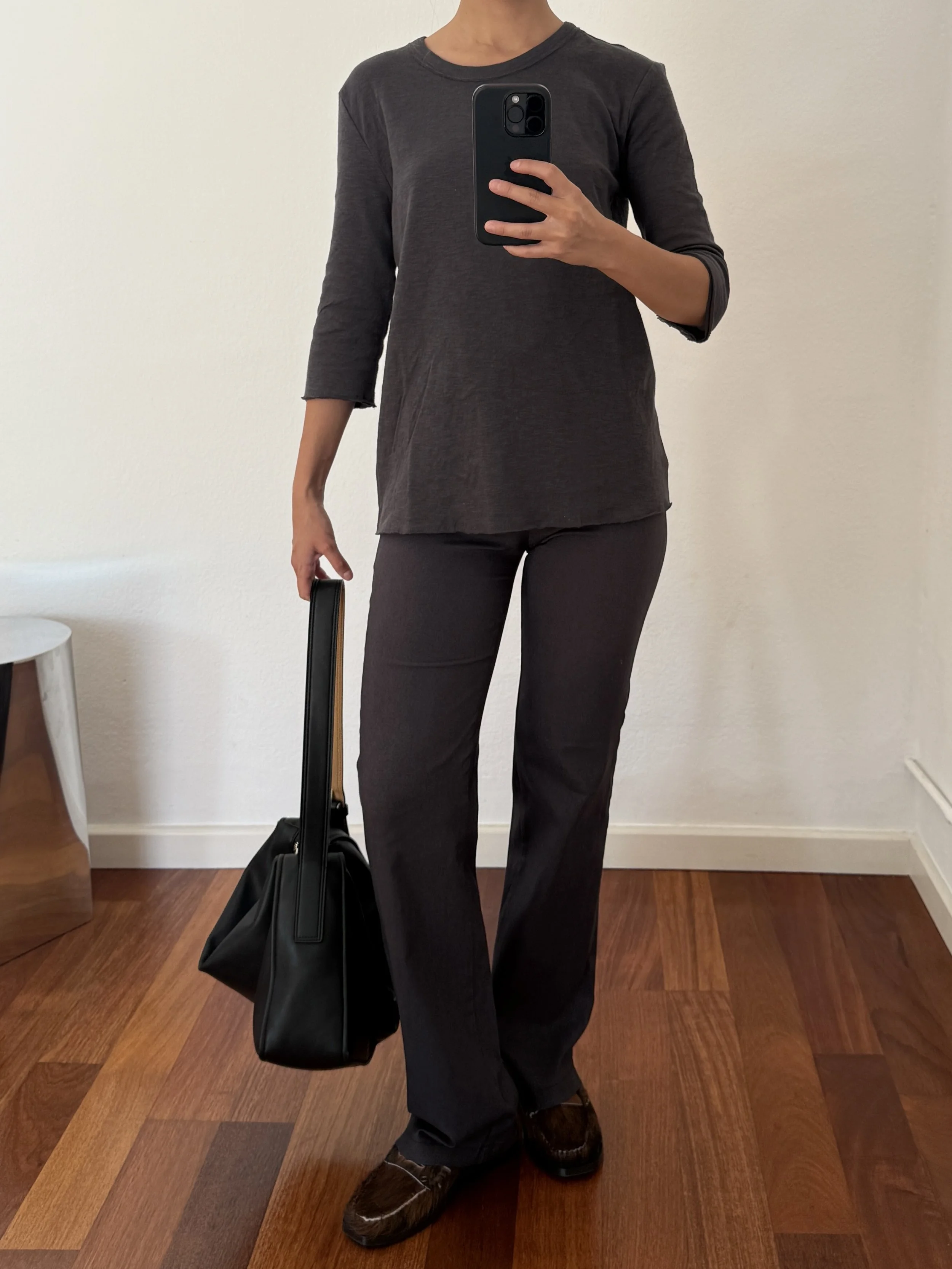 Person taking a mirror selfie wearing a dark gray long-sleeve top and matching dark gray wide-leg pants, holding a black handbag, standing on a wooden floor.