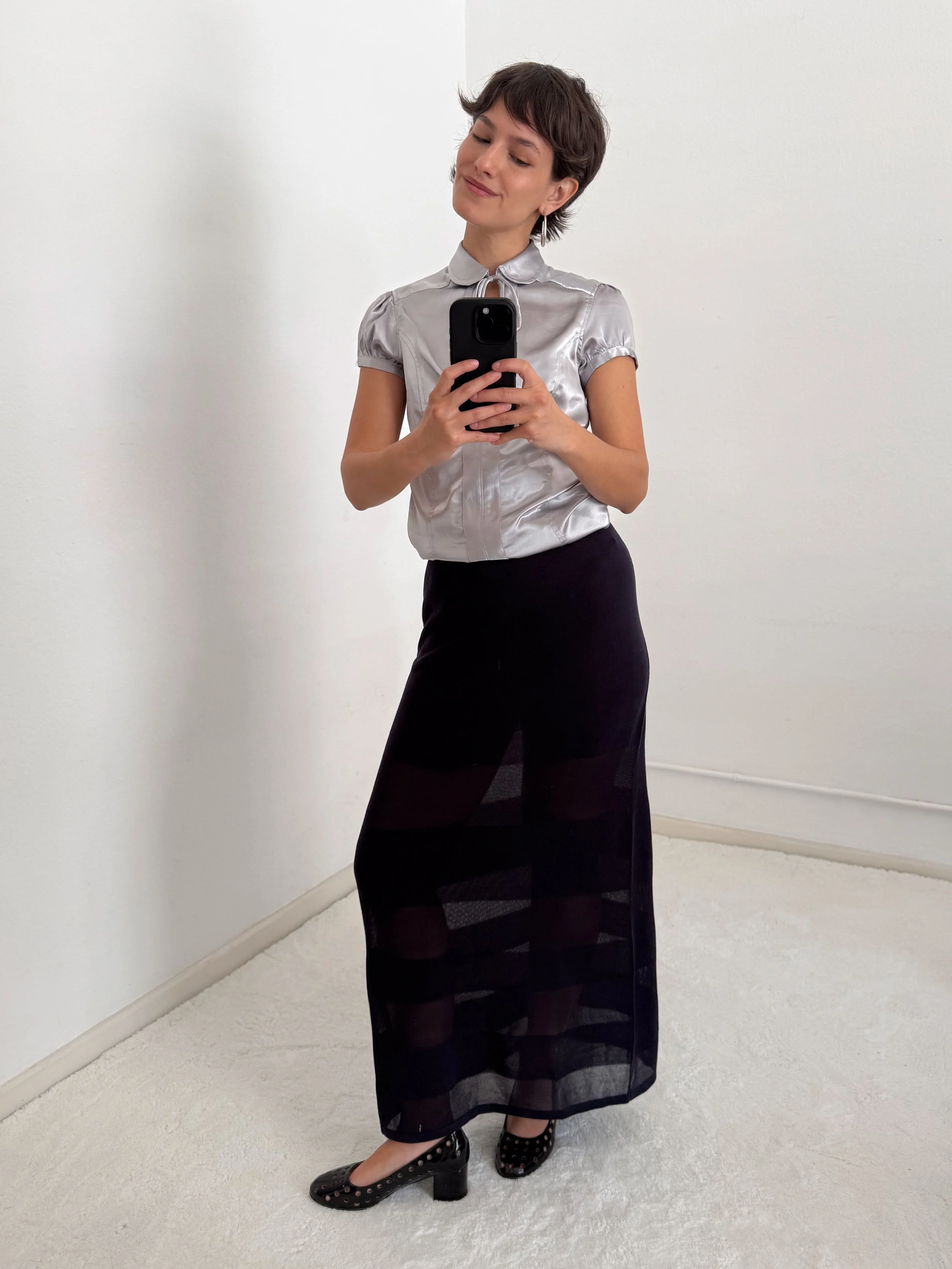 Woman taking a selfie in a mirror, wearing a shiny silver top, a long black skirt with sheer panel, and black shoes with studded design.