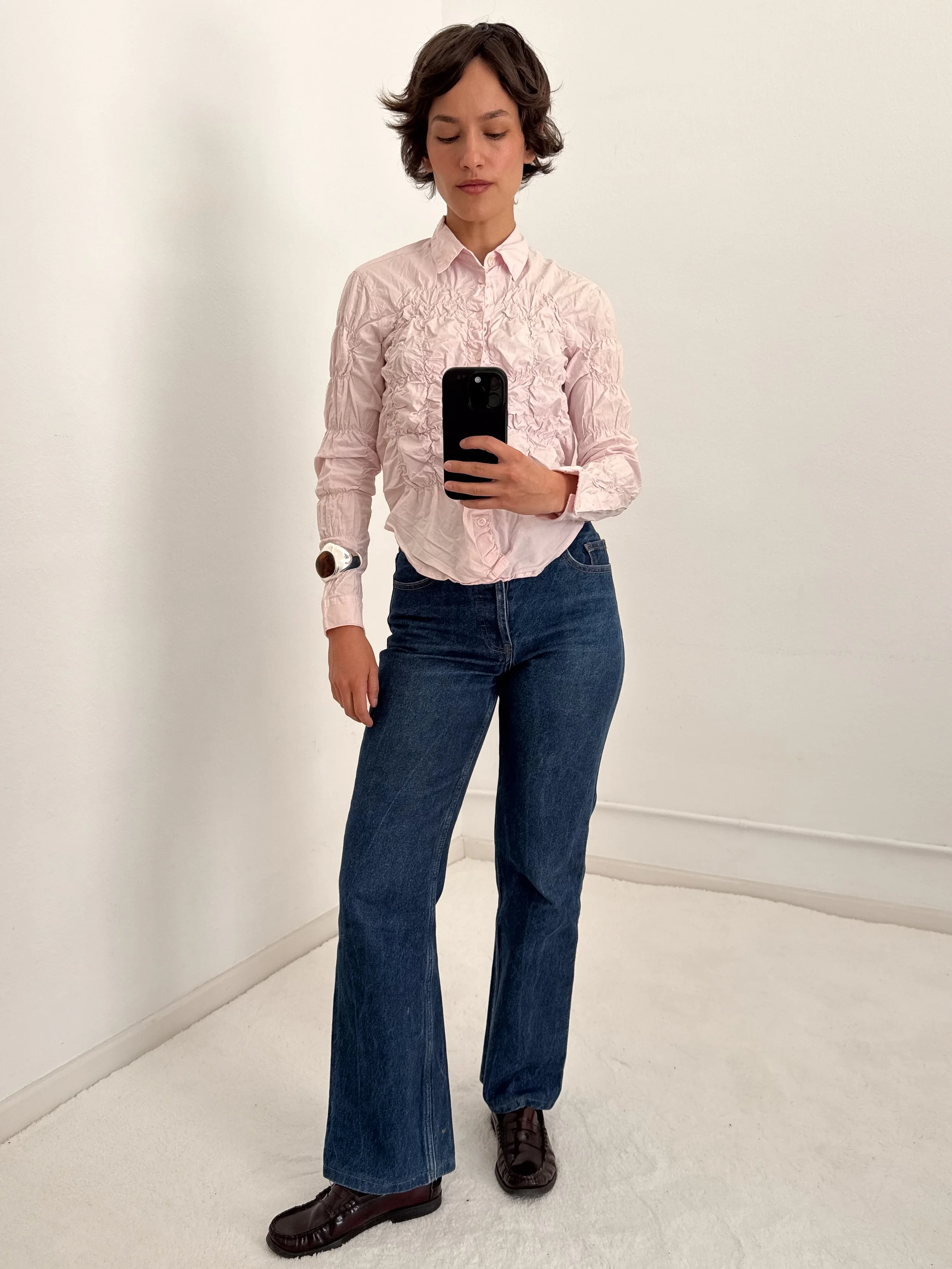 A woman taking a mirror selfie in a plain white room. She is wearing a light pink, crinkly button-up shirt, dark blue wide-leg jeans, and dark brown loafers. She has short, wavy brown hair and is wearing a wristwatch.