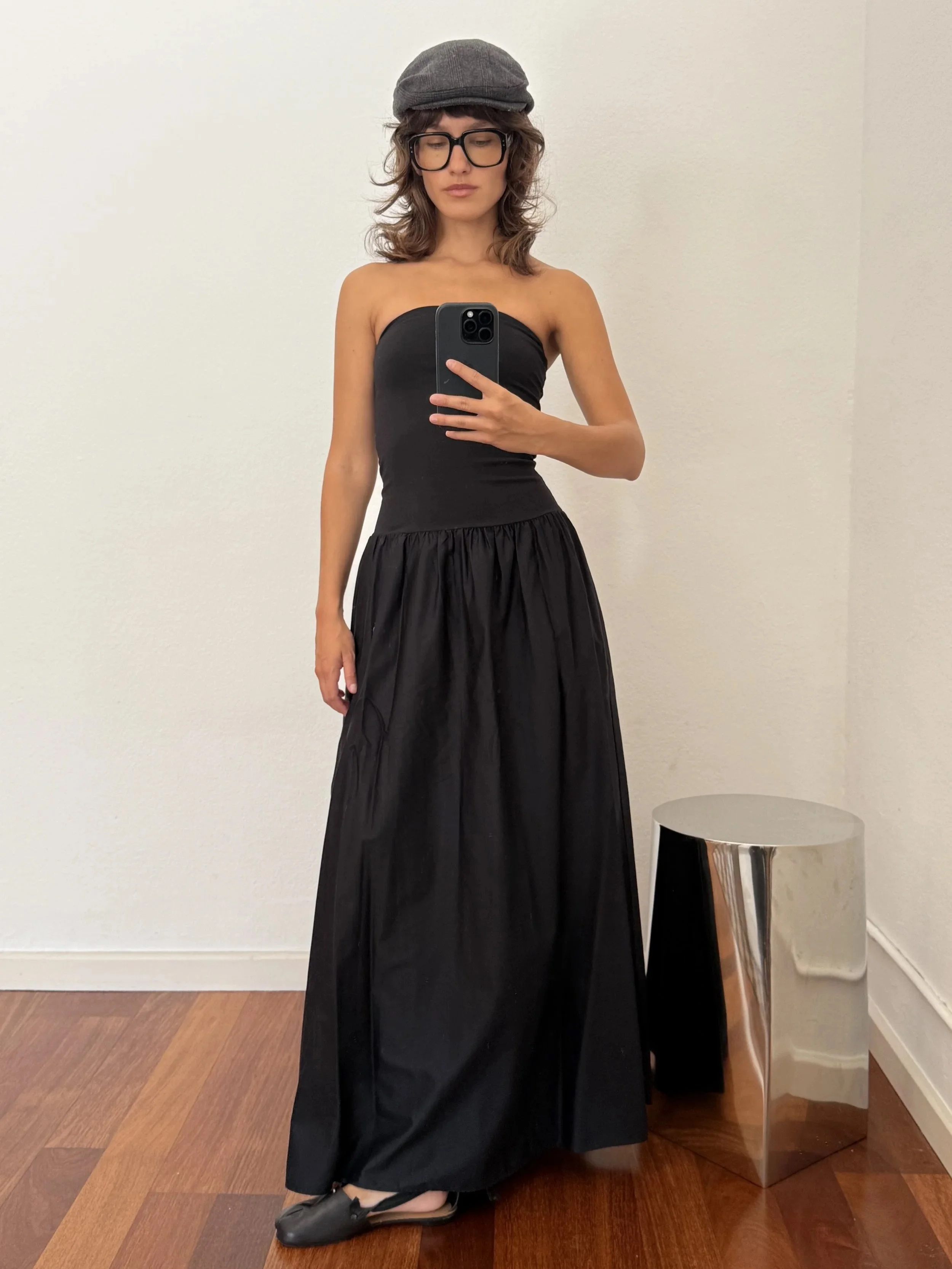 A woman in a strapless black dress and black shoes takes a mirror selfie in a room with hardwood floors. She is wearing glasses and a gray flat cap. There is a round silver side table next to her.