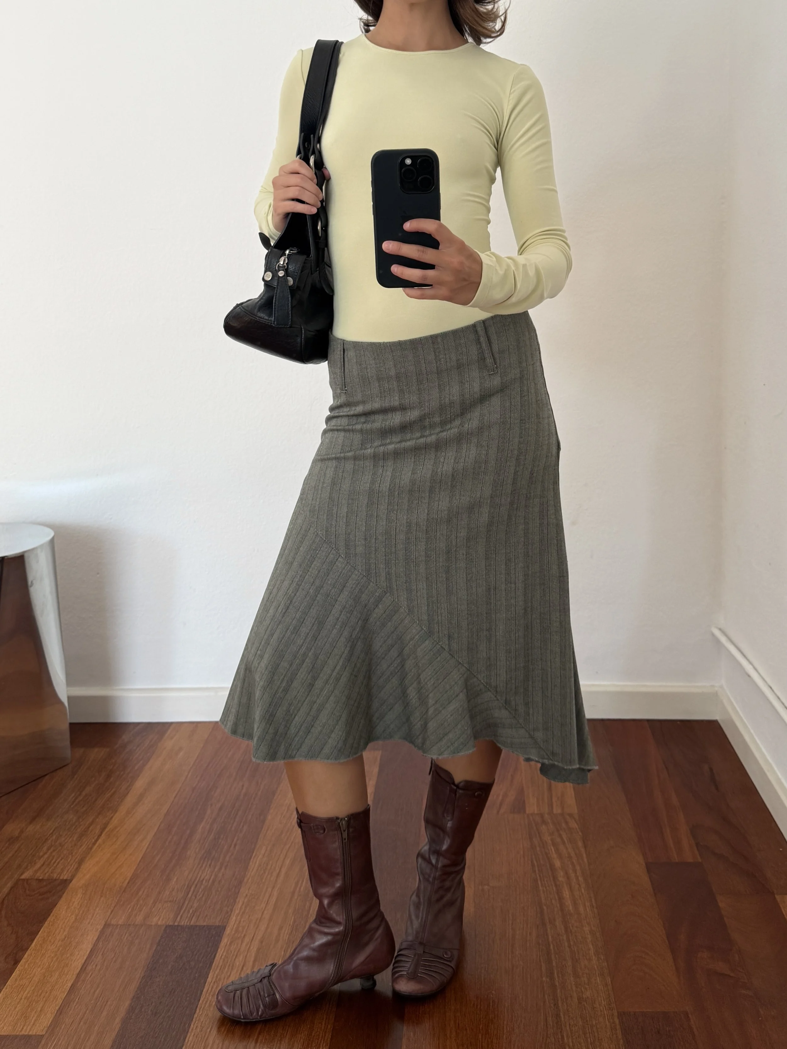 Person taking a mirror selfie wearing a light yellow long-sleeve top, a gray pleated midi skirt, and brown knee-high boots. They are carrying a black shoulder bag.