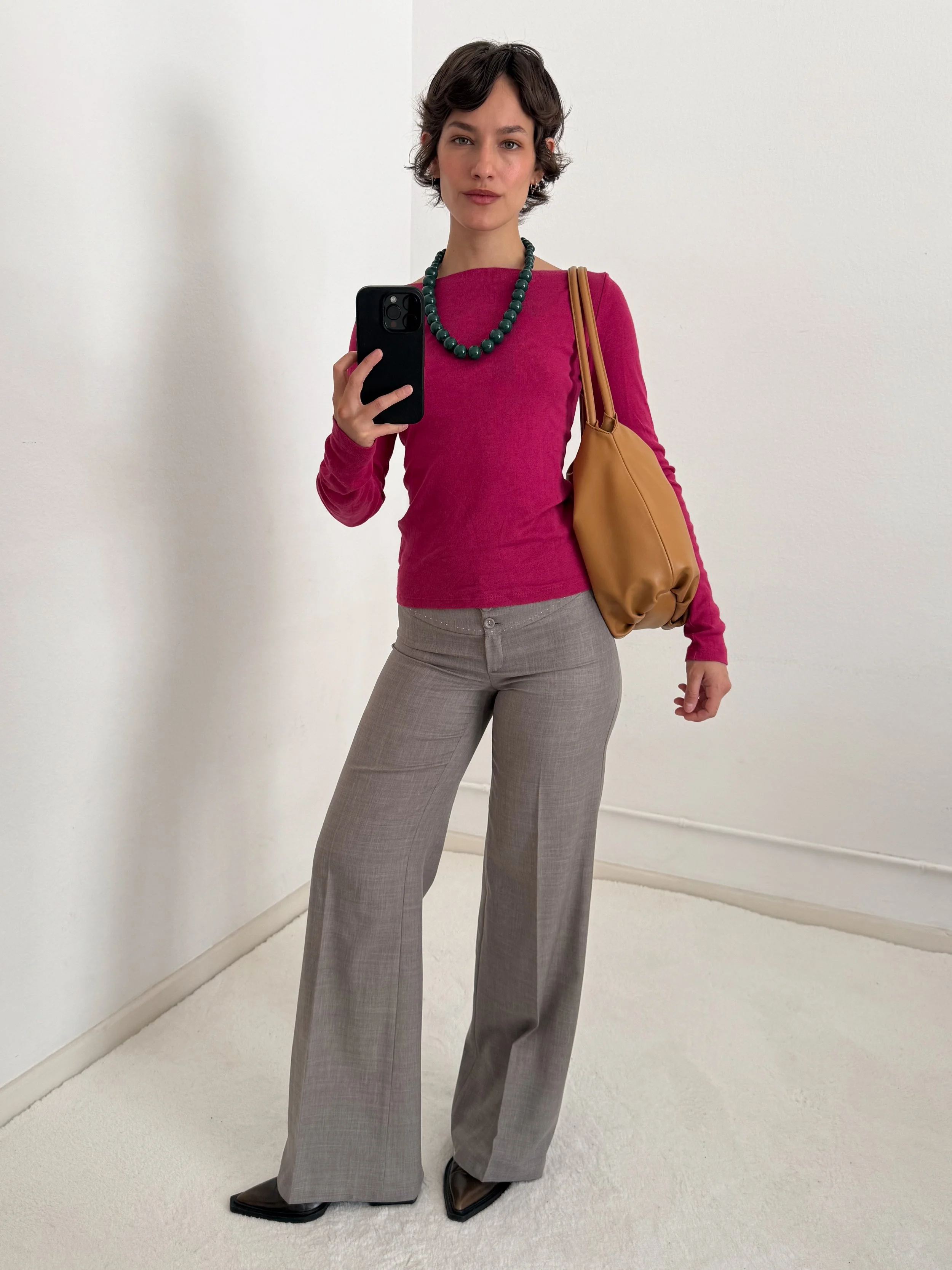 A woman with short dark hair taking a selfie in front of a white wall. She is wearing a pink long-sleeve top, a large dark green beaded necklace, gray wide-leg pants, and black shoes. She has a tan shoulder bag on her left shoulder.