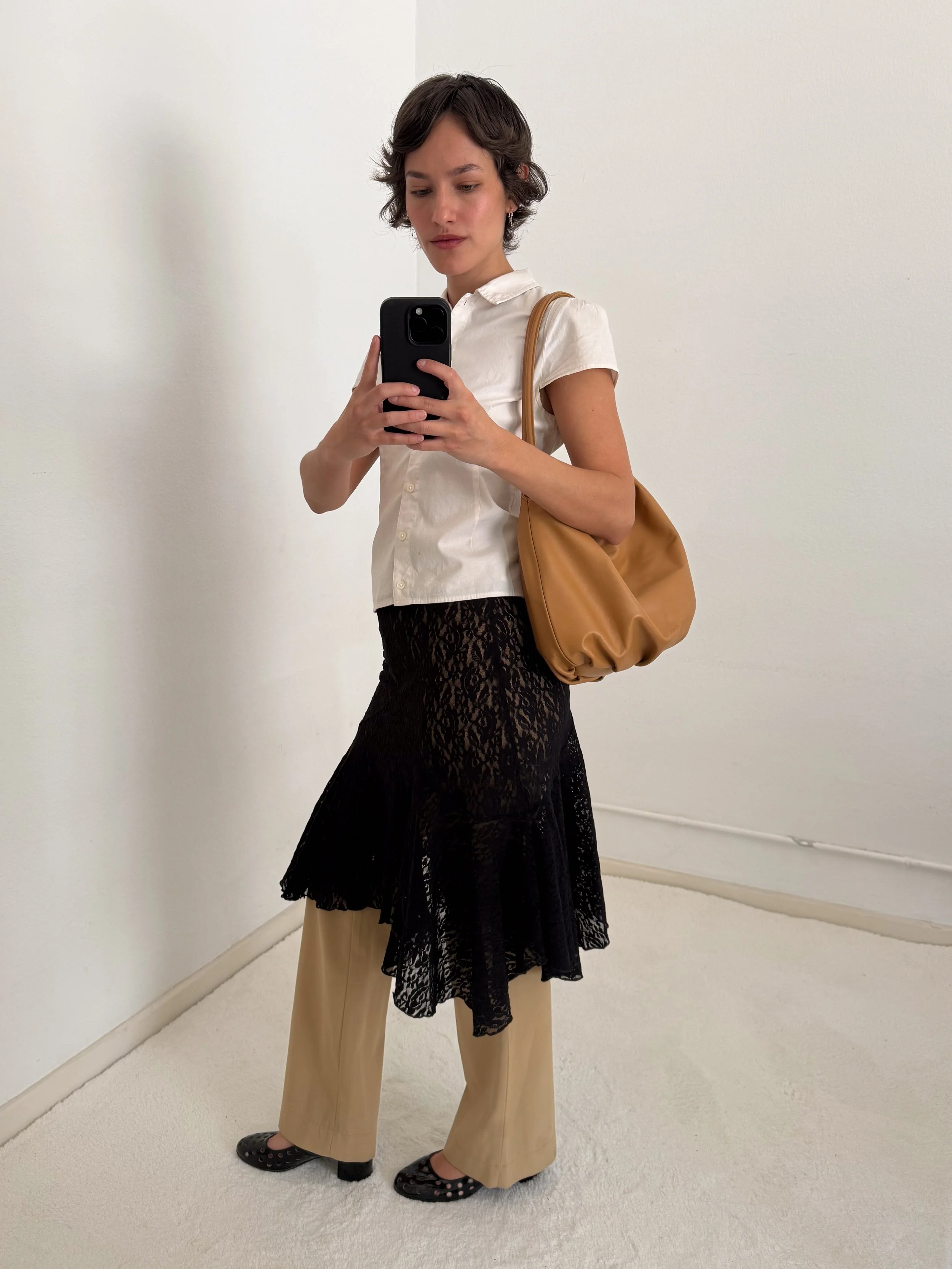 A woman with short wavy hair taking a mirror selfie in a white room. She is wearing a beige wide-leg pants, a white button-up shirt, a black lace skirt layered over the pants, black shoes with cut-out designs, and is carrying a tan shoulder bag.