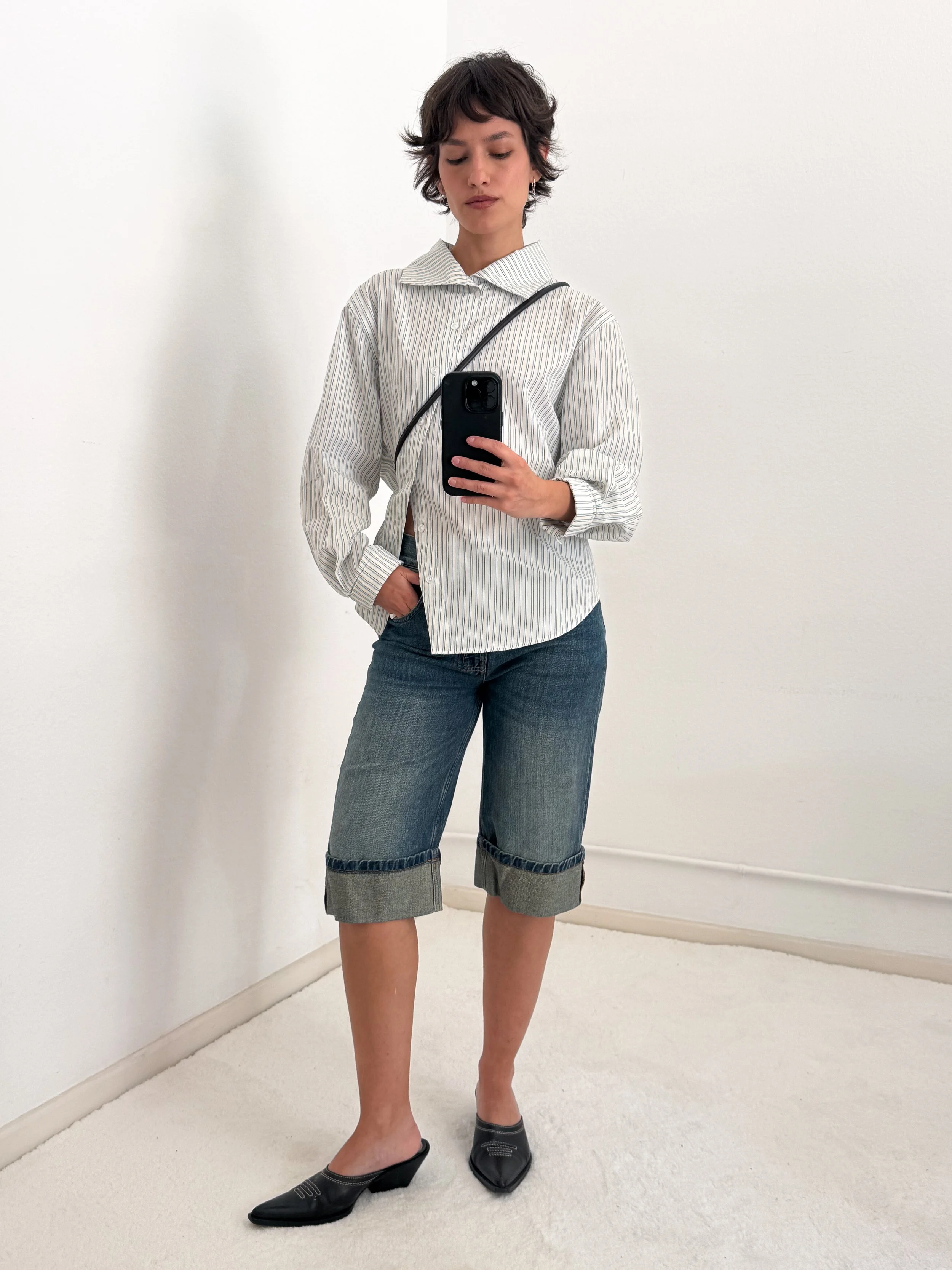 A woman with short curly hair wearing a striped button-up shirt, jeans with rolled-up cuffs, and black heeled shoes, standing against a white wall and taking a selfie with her phone.