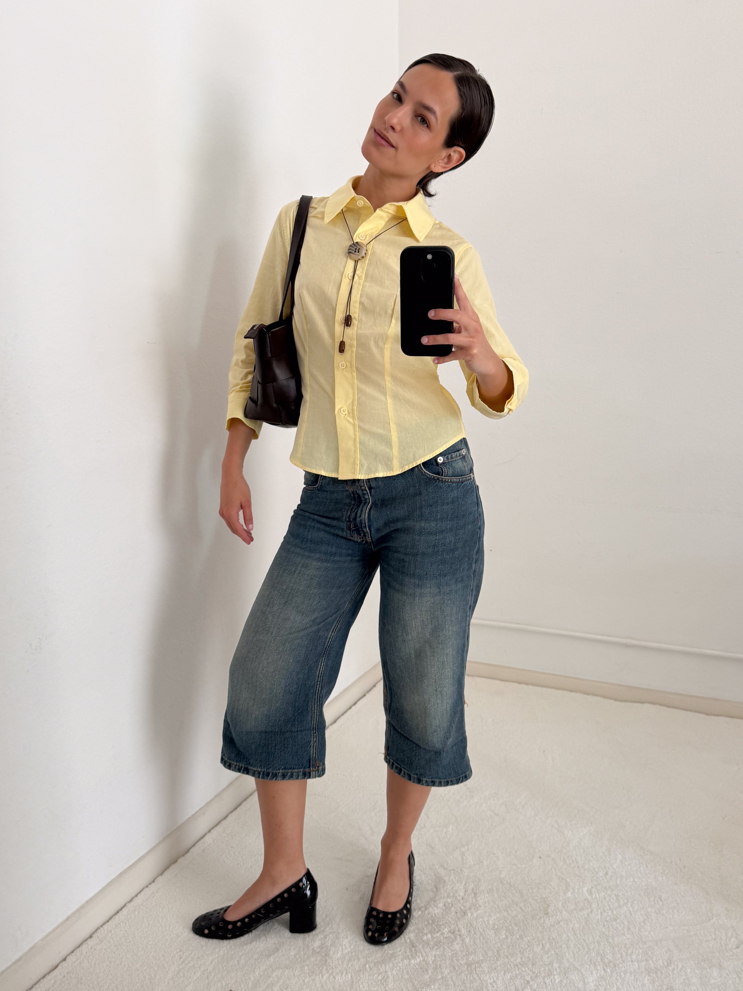 Woman taking a mirror selfie, wearing a yellow button-up shirt, dark jeans, black studded high heels, and carrying a black shoulder bag, standing against a white wall.